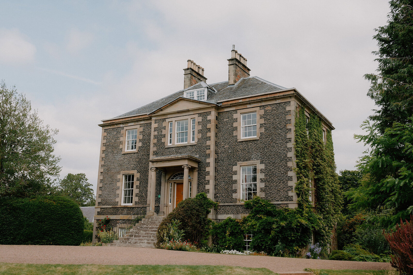 A stone country house with a pillared entrance porch stands behind a lawn, ivy climbing one corner and mature trees framing the view.