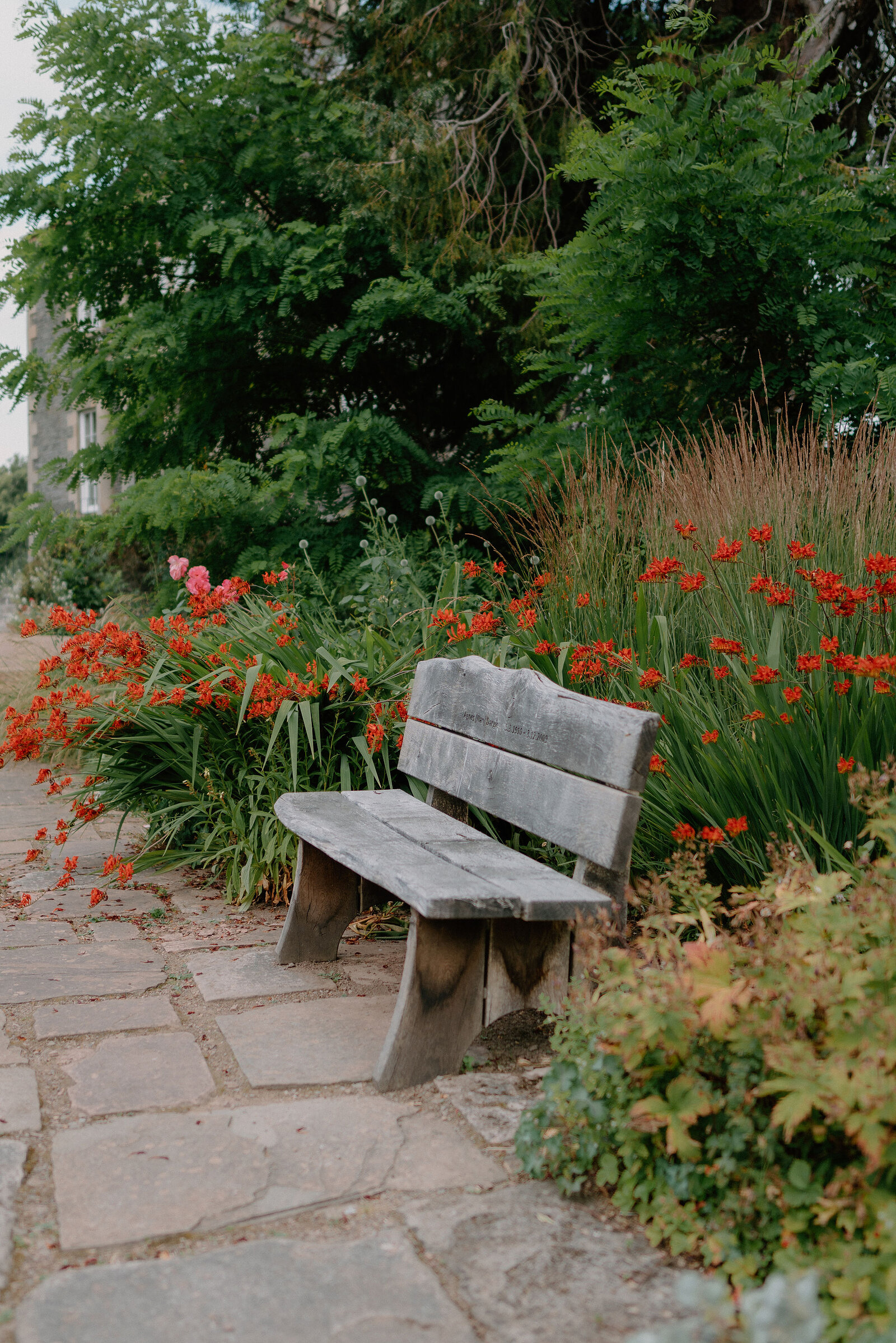 A weathered wooden bench stands on a stone path beside a bed of red crocosmia flowers, mature trees rising behind.