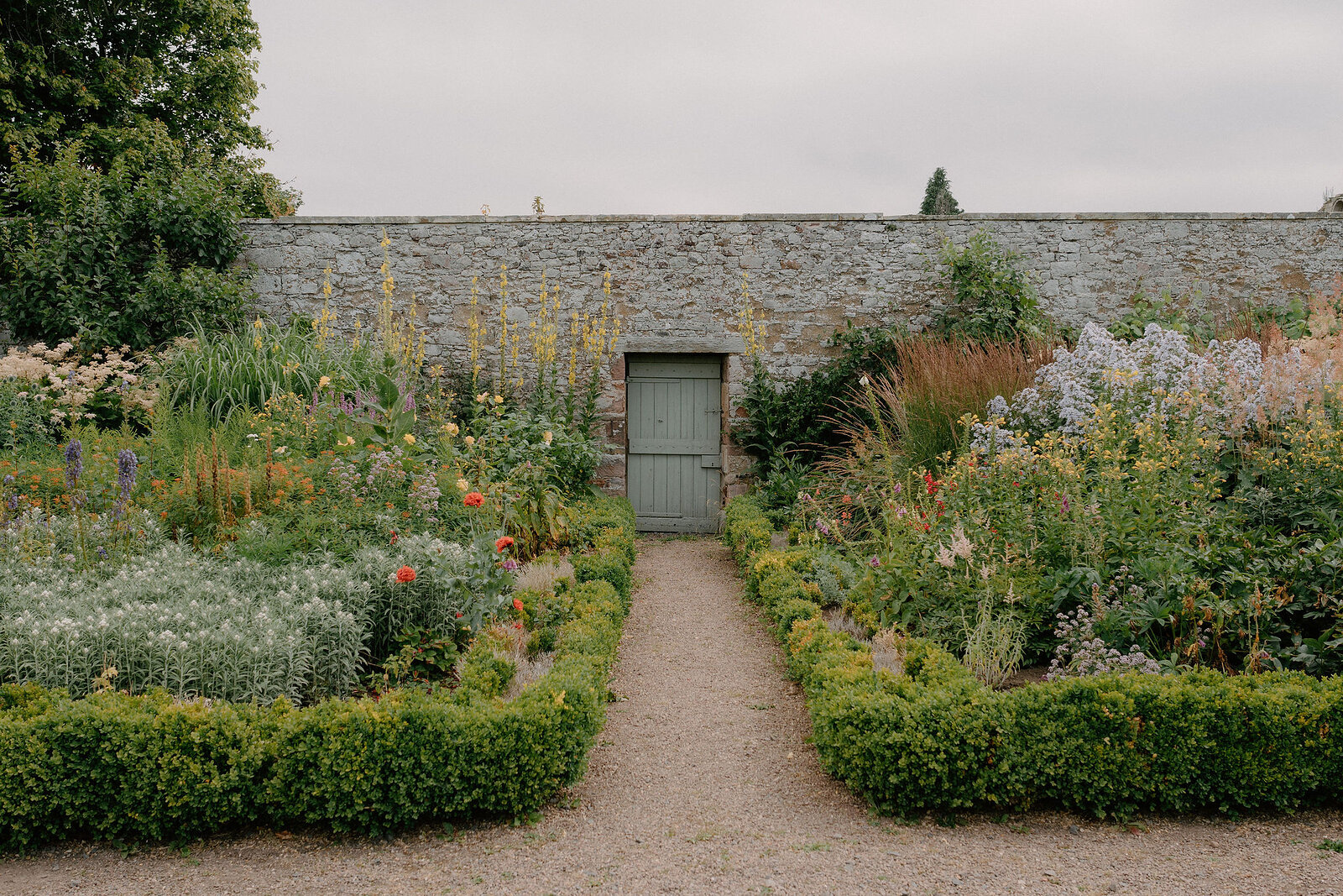 A wooden door in a stone garden wall stands framed by tall flowering borders, a gravel path leading between clipped box hedges.