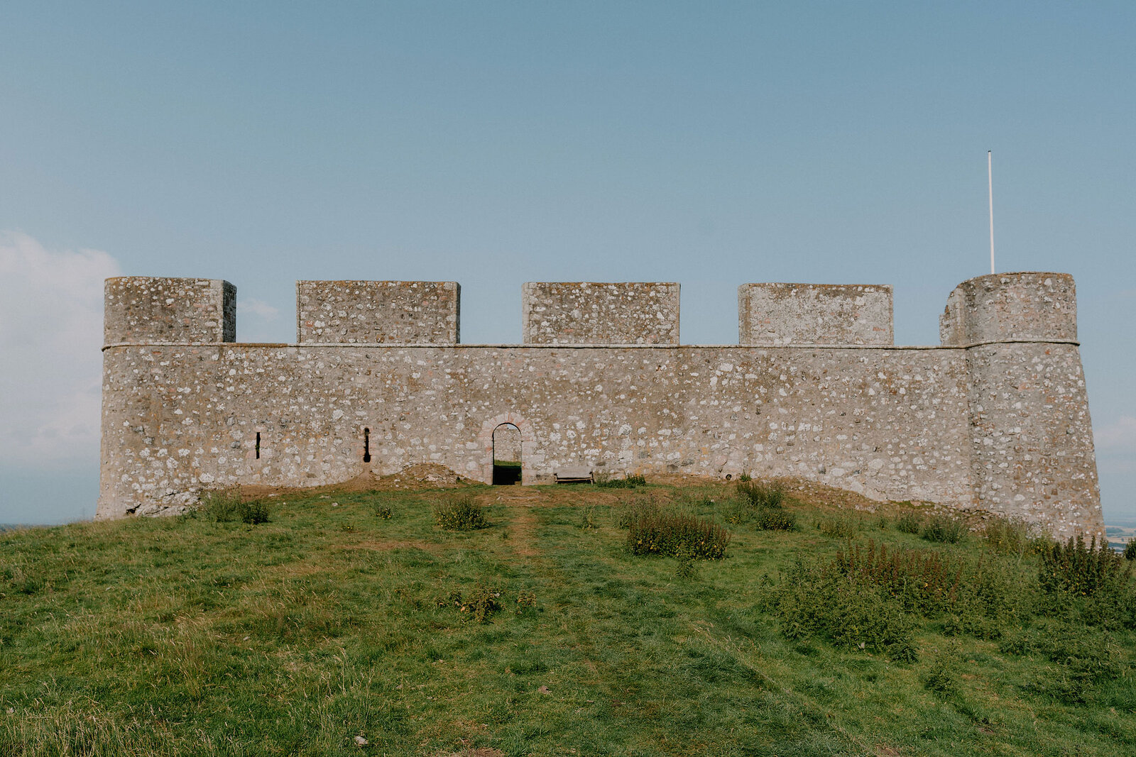 A crenellated stone fort with round corner towers stands on a grassy mound, a flagpole rising from one corner and a plain doorway set into the wall.
