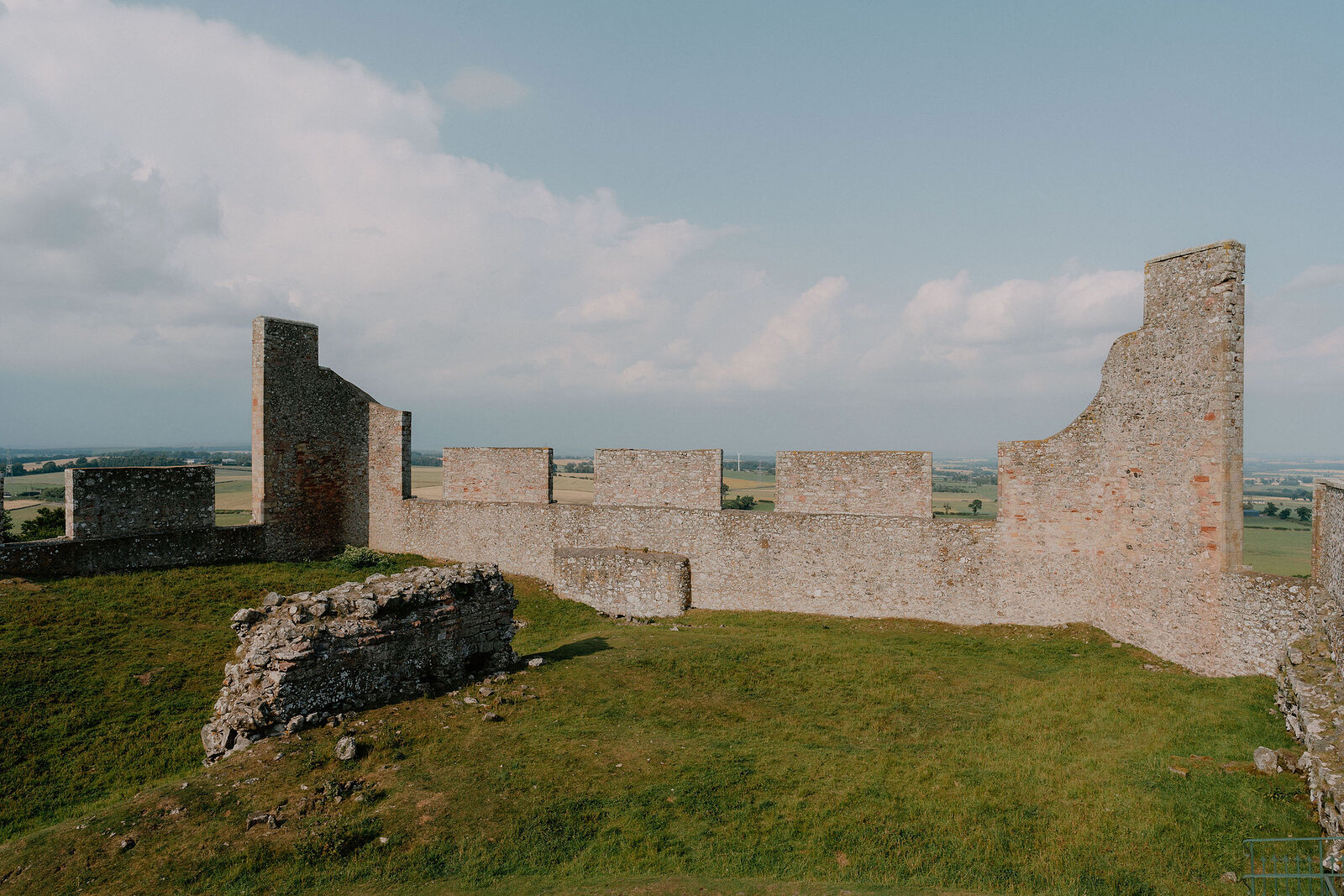The battlemented walls of a ruined fort stretch across a grassy headland, open fields visible beyond the crumbling stonework.