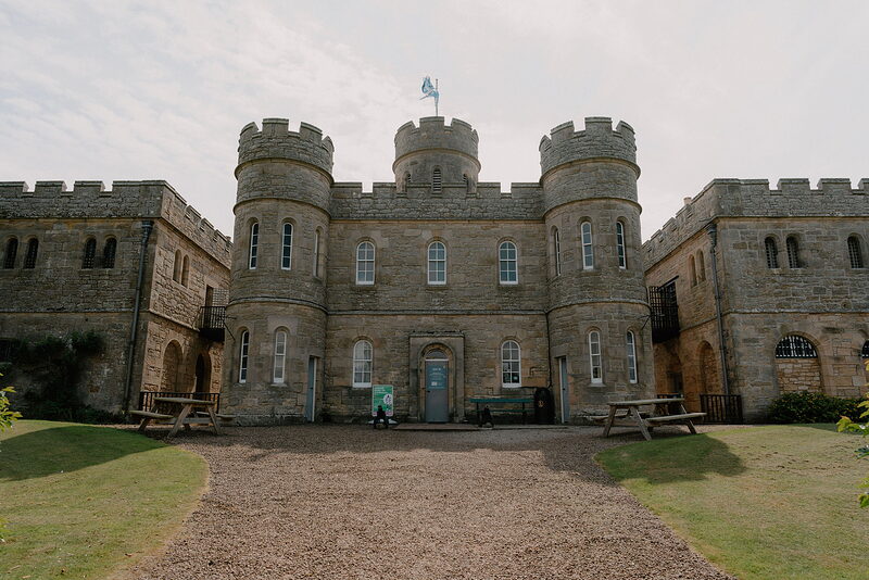 A crenellated stone fort with corner towers rises behind a gravel forecourt, picnic tables set on the lawn to either side.