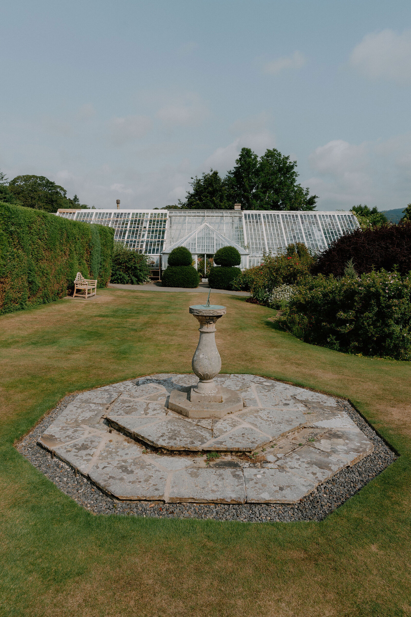 A stone sundial stands on an octagonal paved platform in a lawned garden, a long glasshouse and clipped topiary behind it and a wooden bench to one side.