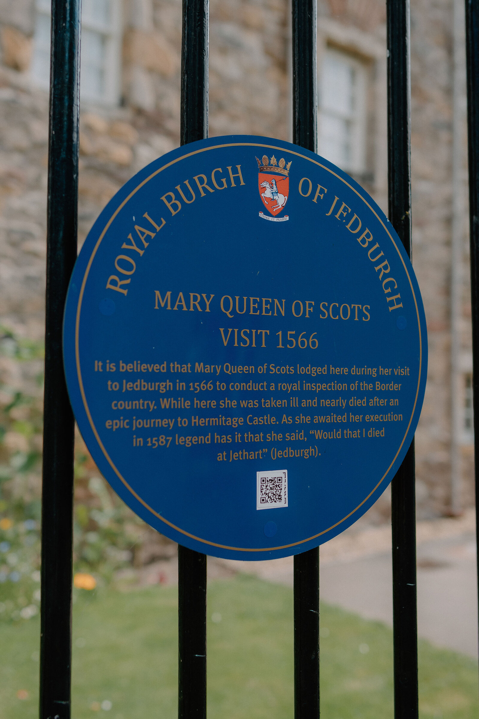 A blue plaque reading "Royal Burgh of Jedburgh, Mary Queen of Scots visit 1566" is mounted on iron railings in front of a stone building.
