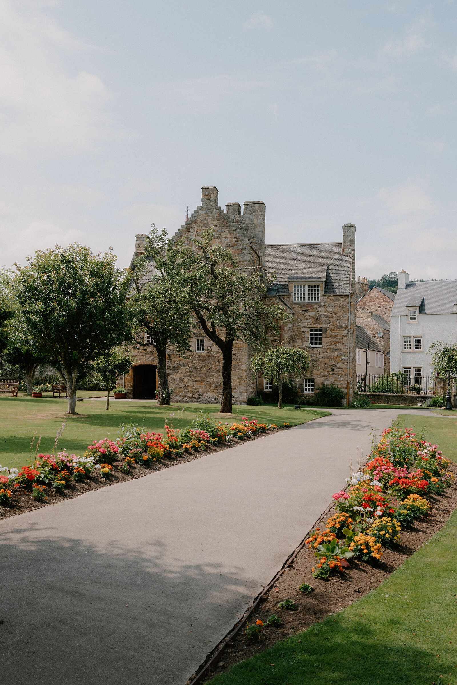 A stone tower house with crow-stepped gables stands beyond a lawn dotted with trees, a bench nearby.