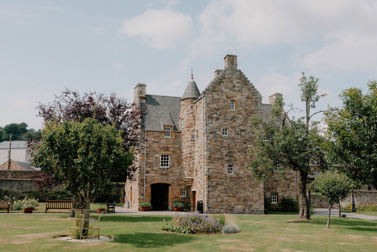 A stone tower house with a turret and crow-stepped gables stands beyond a lawn with benches and flower beds.