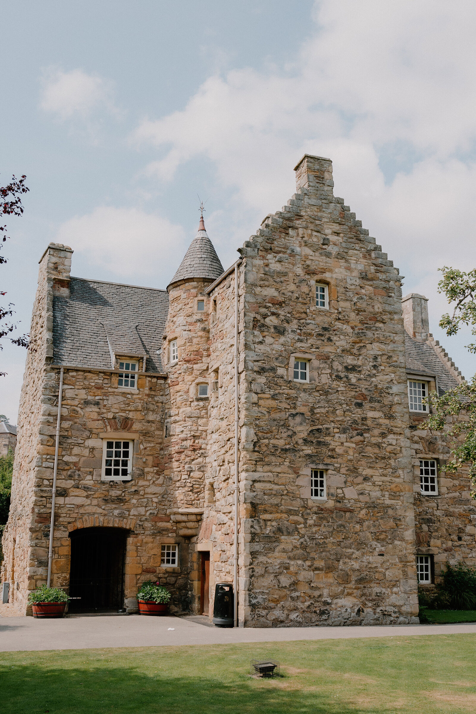 A stone tower house with a turret and an arched entrance stands beside a lawn, potted plants at its base.