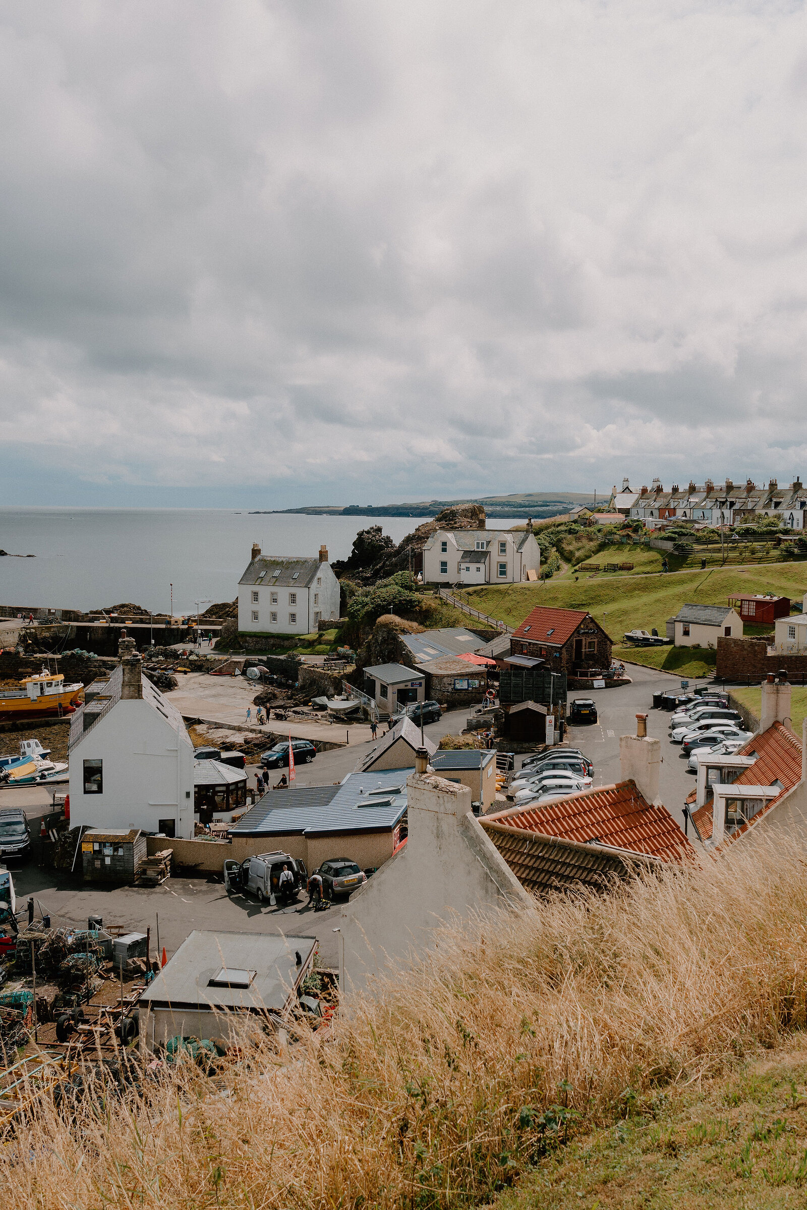 An aerial view shows a fishing village of white and stone houses clustered around a harbour, cars parked along the quayside and cliffs rising behind.