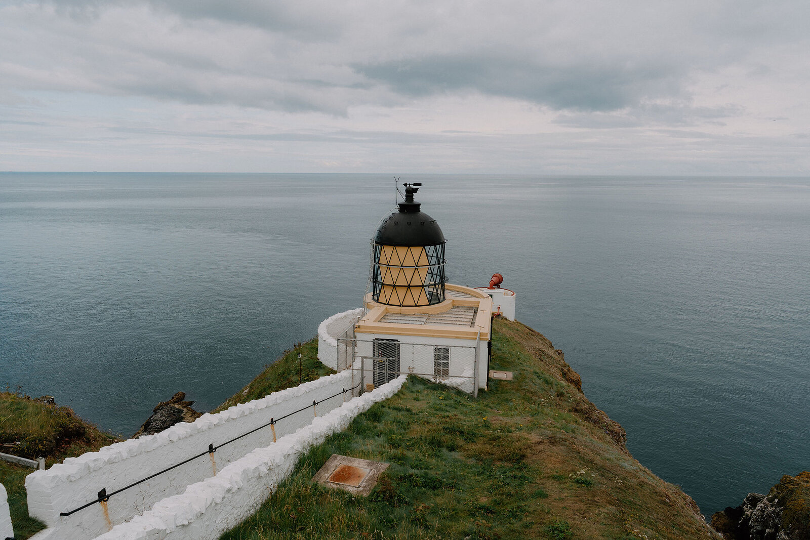 A lighthouse with a black and yellow lantern room stands at the tip of a grassy clifftop, whitewashed walls lining the path below.