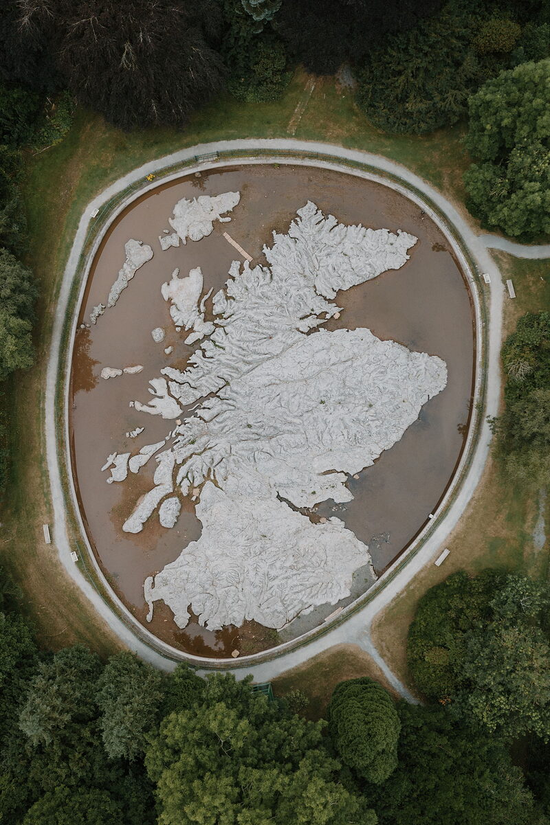 An aerial view shows a huge relief map of Scotland's coastline and islands set into a circular pool, gravel paths and trees encircling it.