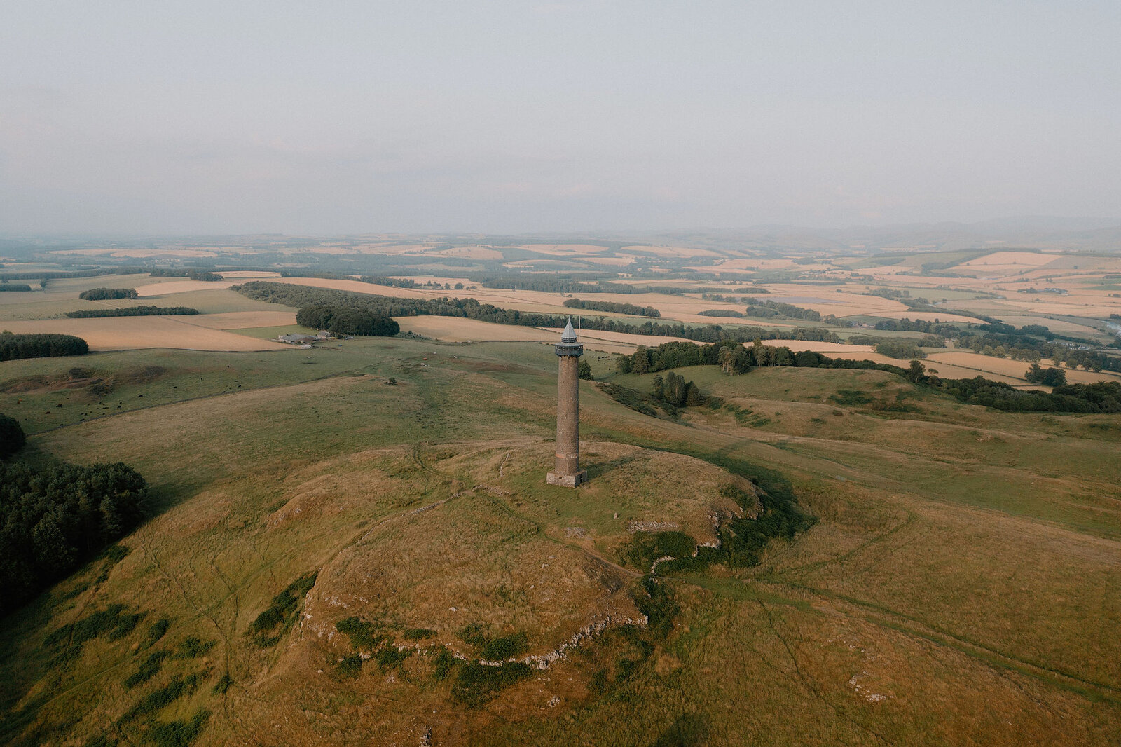 An aerial view shows a tall stone monument standing on a hilltop, farmland and woodland stretching into the hazy distance.