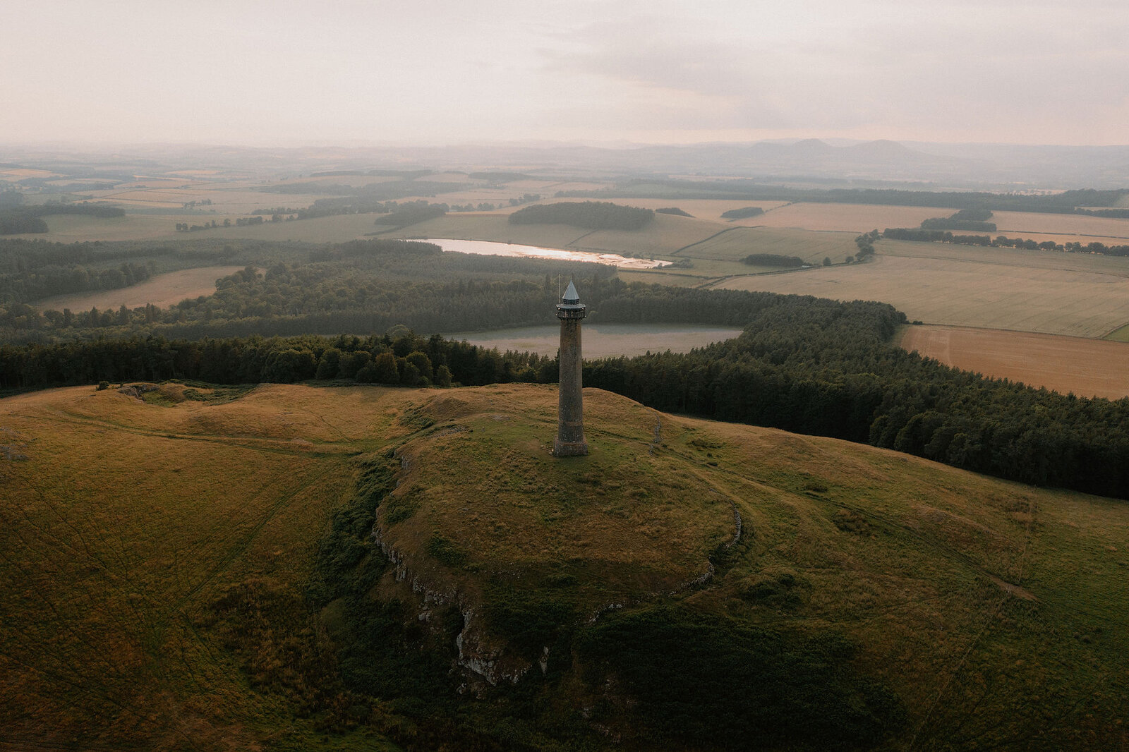 An aerial view shows a tall stone monument standing on a hilltop above a winding river, farmland and woodland stretching into the hazy distance.