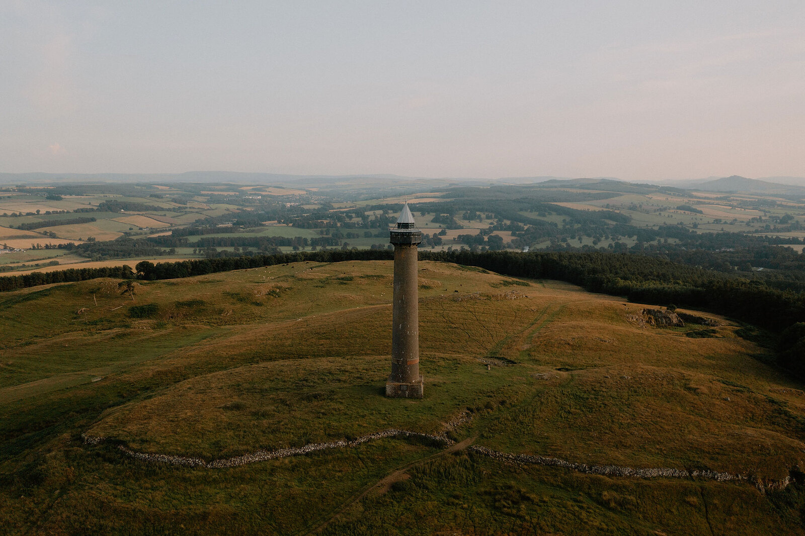 An aerial view shows a tall stone monument standing on a hilltop, farmland and woodland stretching into the hazy distance under a golden evening light.
