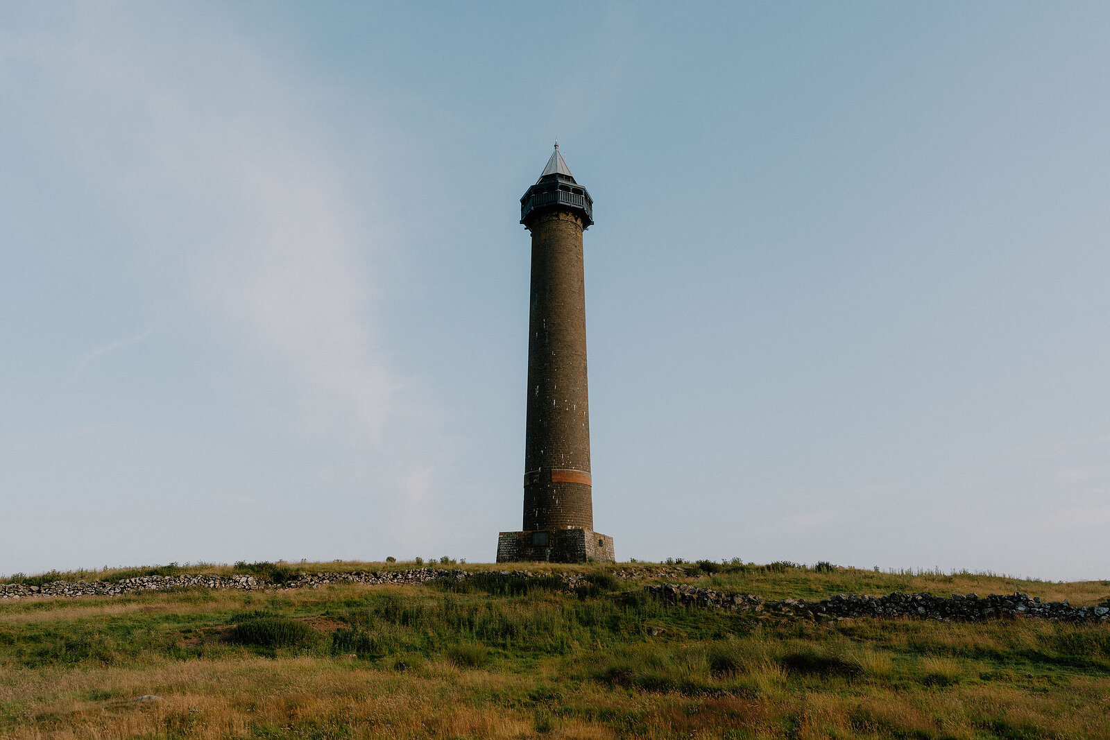 A tall stone monument stands alone on a grassy hilltop bordered by a drystone wall, a clear sky above.