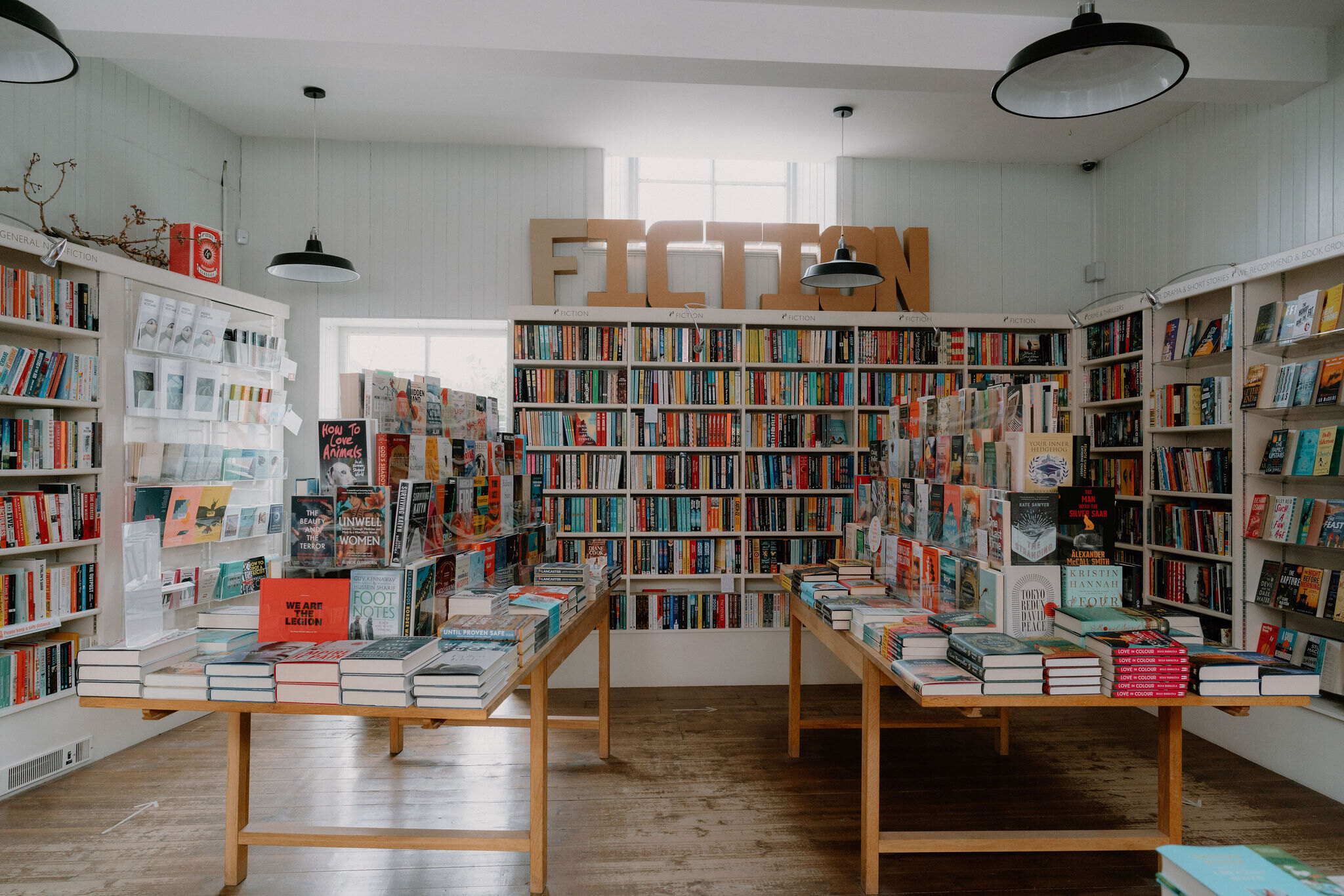 A wooden "Fiction" sign hangs above bookshelves in a bright bookshop, tables piled with books filling the room below pendant lights.