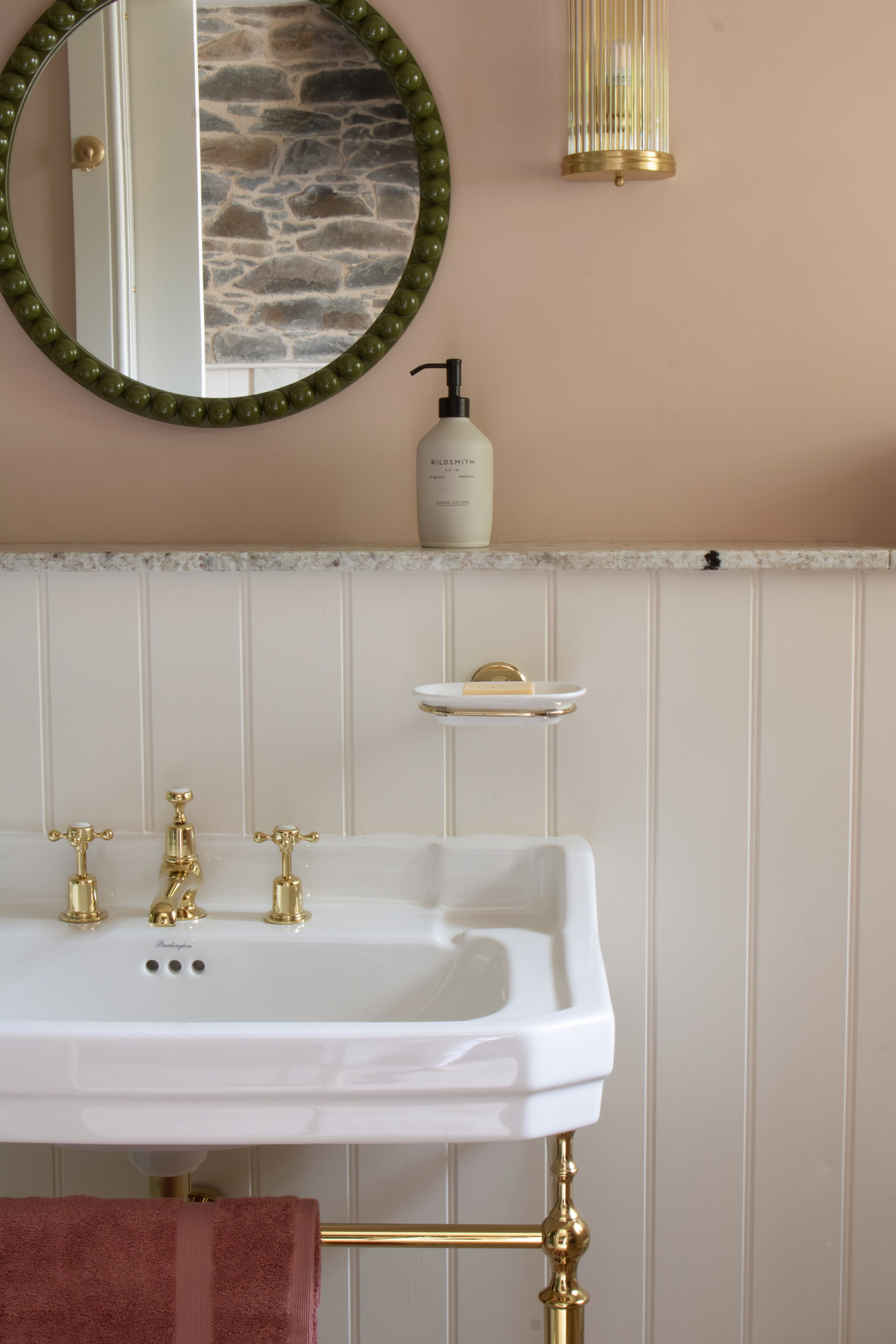A white period basin on brass legs with brass taps, a round beaded mirror on the pink wall above the panelling.