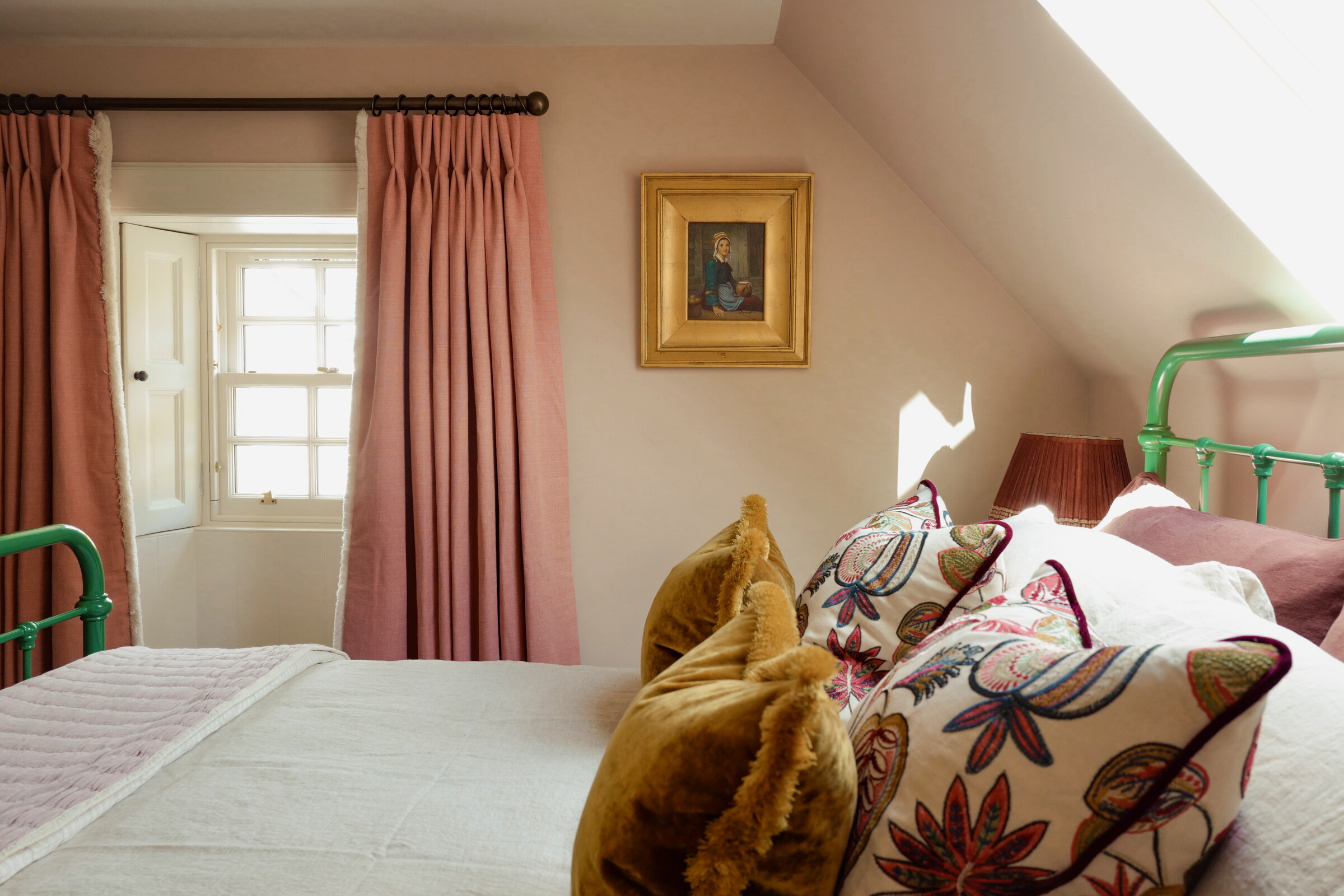 The bed under the sloping ceiling, piled with velvet and embroidered cushions, pink curtains at the window and a portrait in a gilt frame.