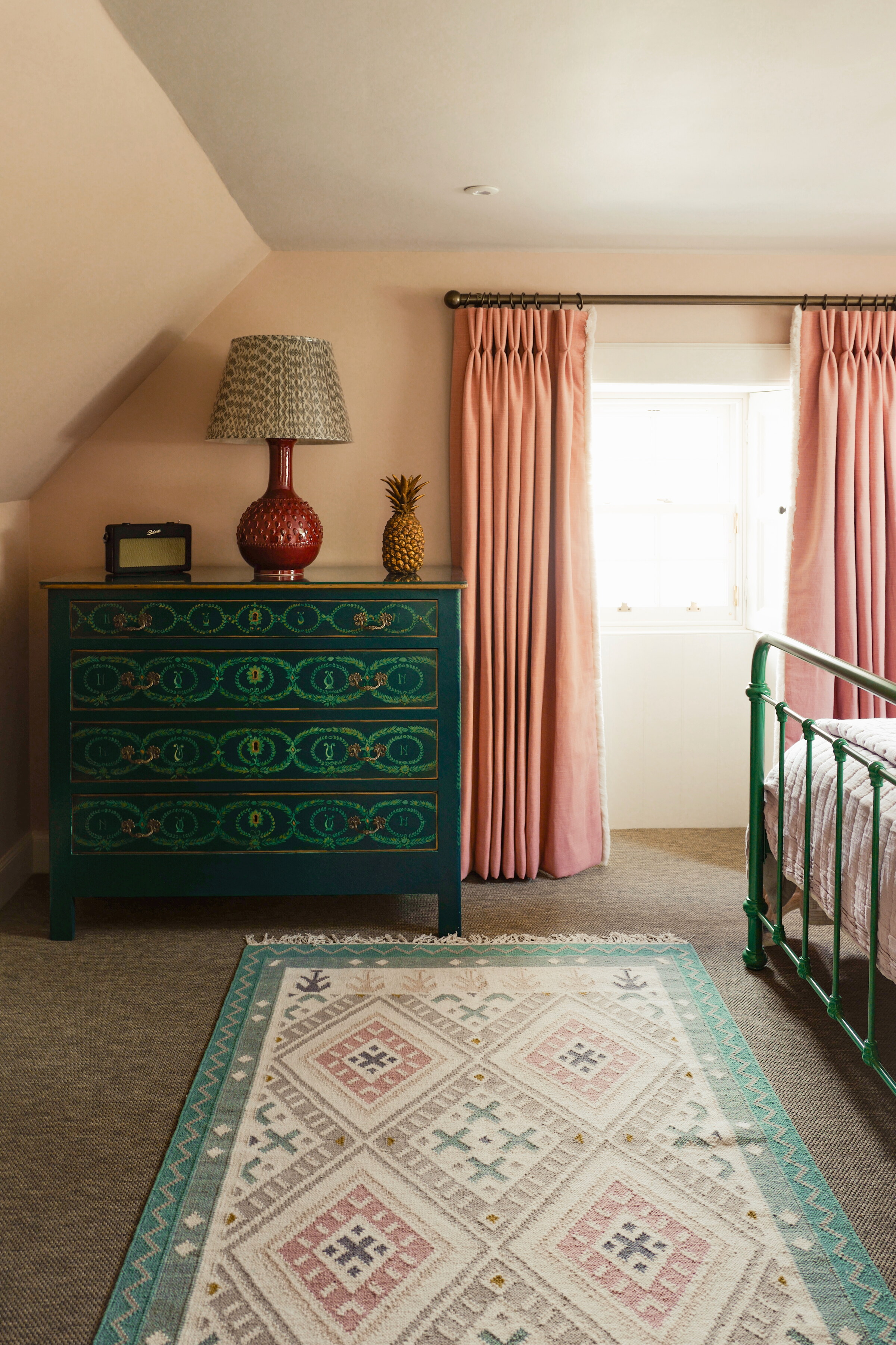 The bedroom under the eaves, a green iron bedstead, a painted chest, a pink and green rug and pink curtains at the window.