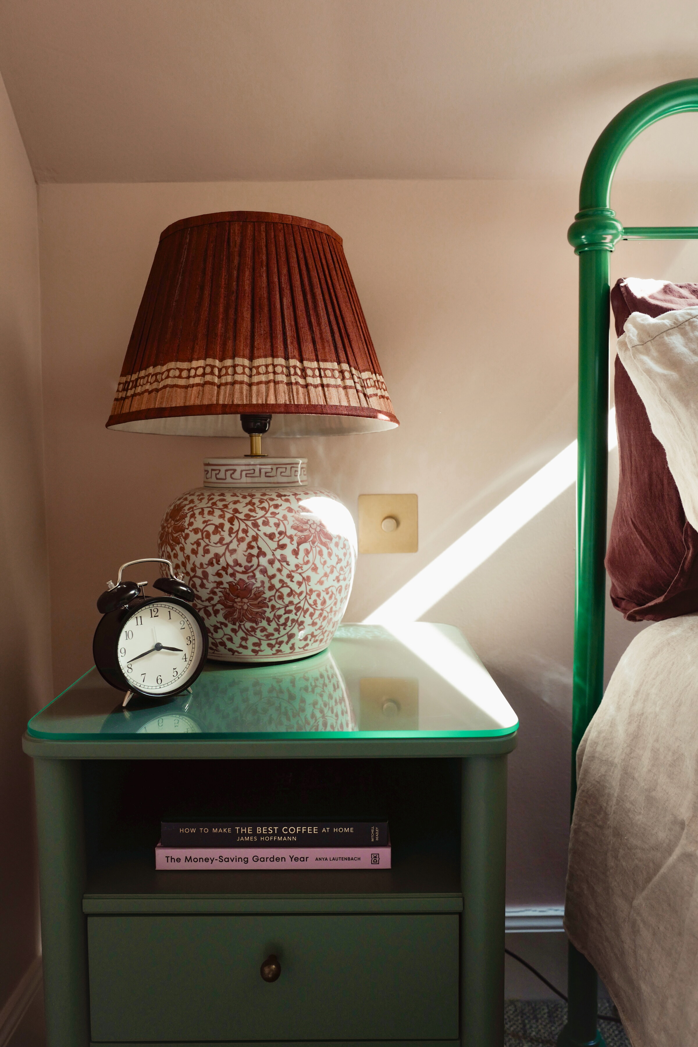 A green bedside table with an alarm clock and a pleated-shade lamp beside the green iron bedstead.