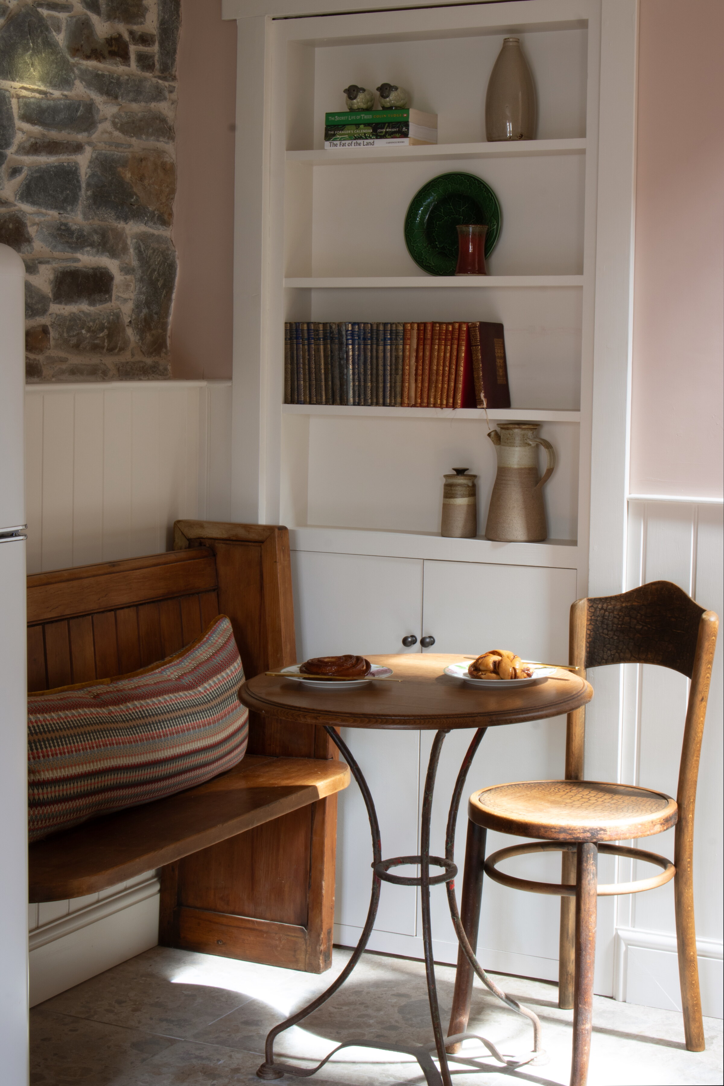 A pew and a bentwood chair at a small round table laid with pastries, beside white shelves of books and pottery.