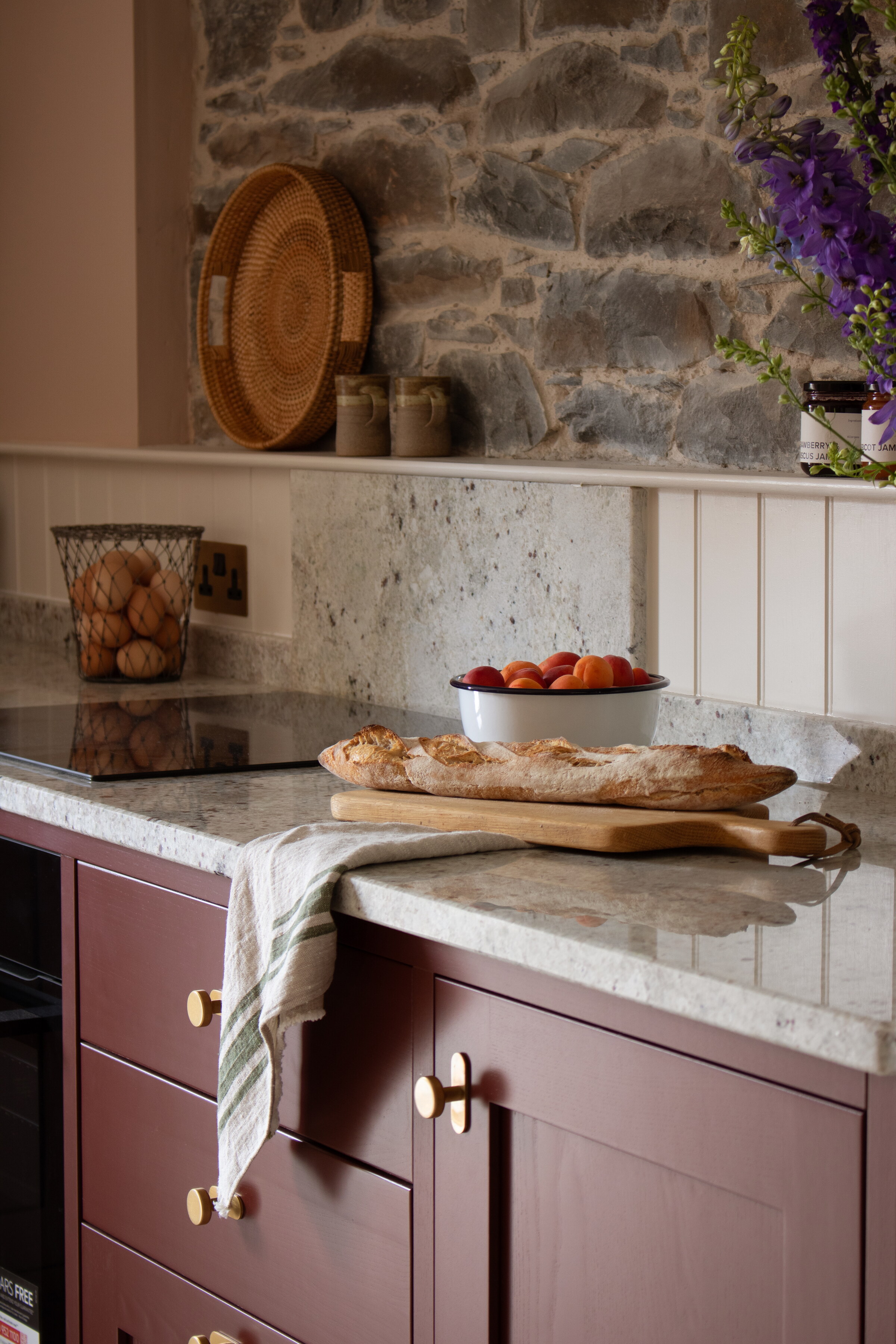 A loaf on a board and a bowl of tomatoes on the speckled granite worktop above plum cupboards with brass knobs.