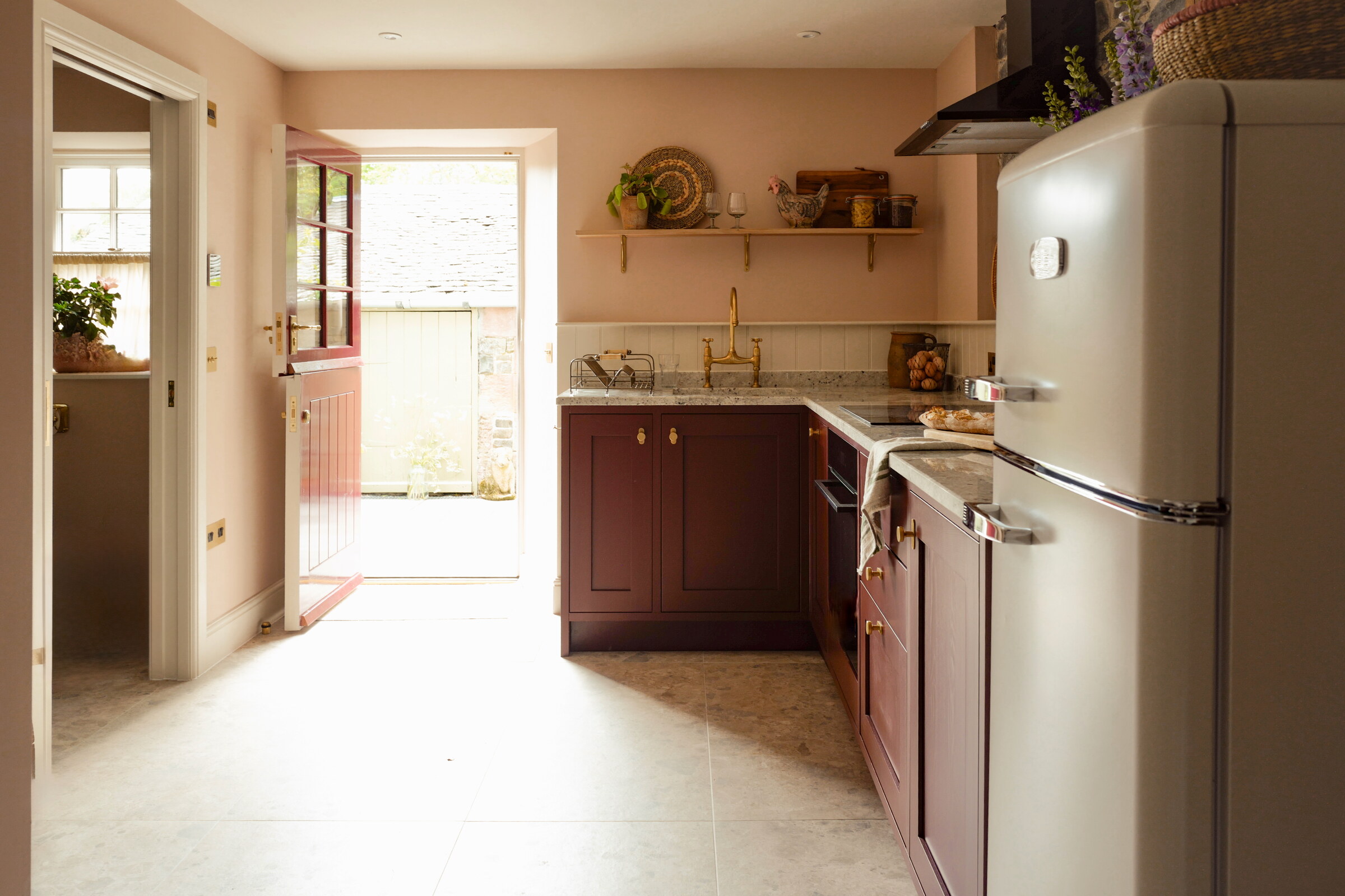 The kitchen from the hall, plum cupboards and a cream fridge, the stable door open to the courtyard at the end.