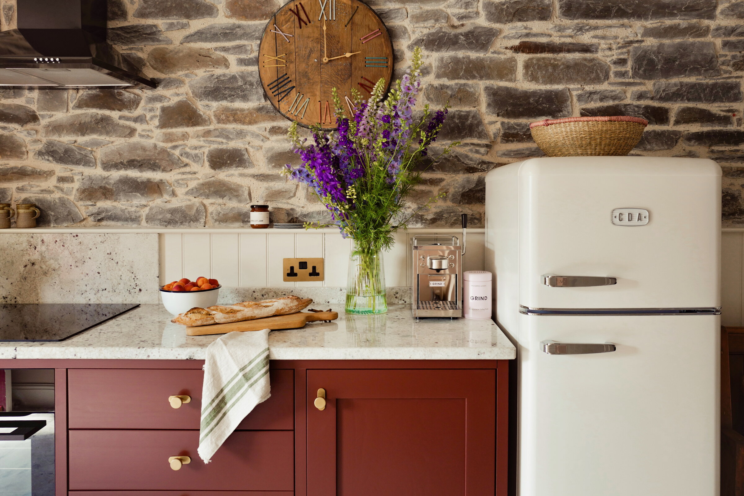 The kitchen’s plum cupboards and pale granite worktop under an exposed stone wall, with a cream fridge, a clock and a jug of delphiniums.
