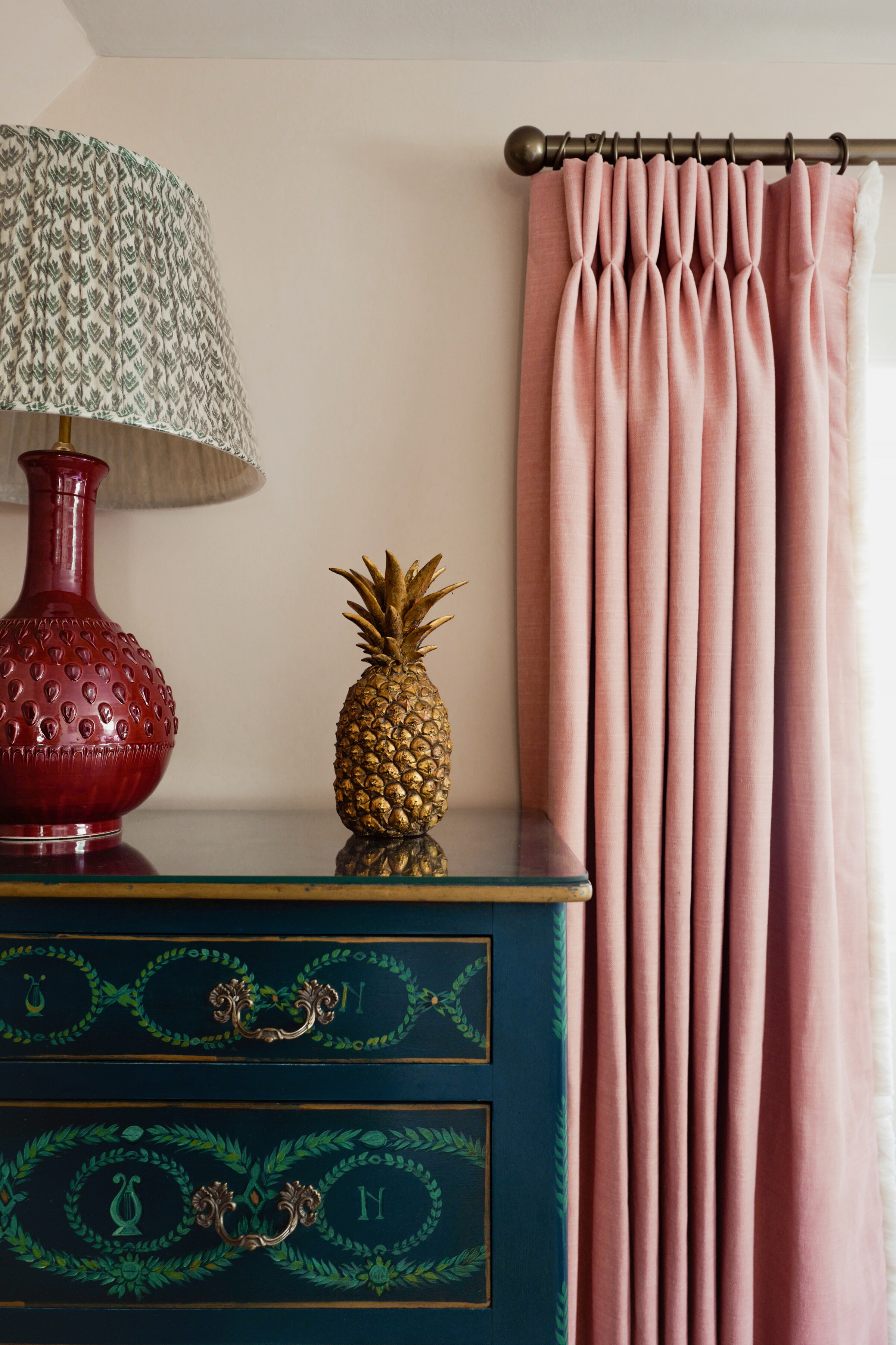 A painted green and gold chest of drawers with a red lamp and a gilded pineapple, pink pleated curtains beside it.