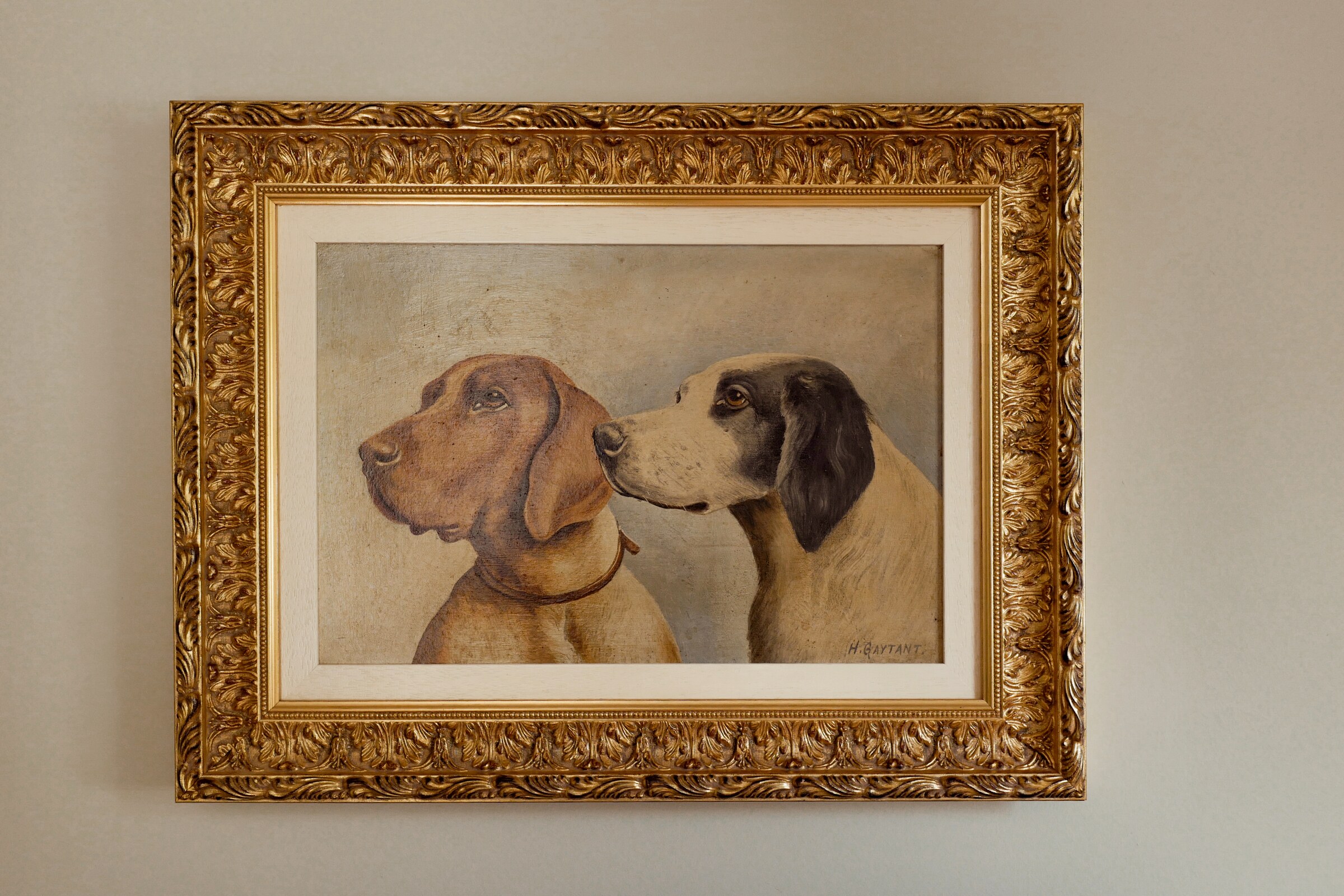 An old oil painting of two hounds’ heads in an ornate gilt frame on a pale wall.
