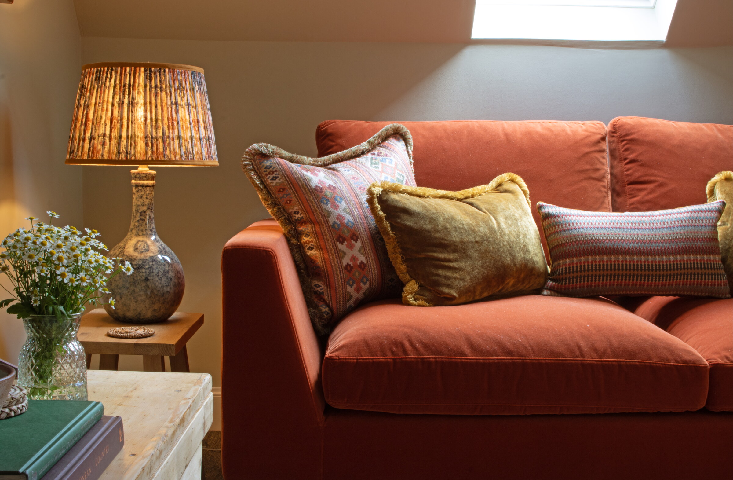A rust velvet sofa heaped with patterned and gold cushions beside a lamp with a pleated shade and a jar of daisies.