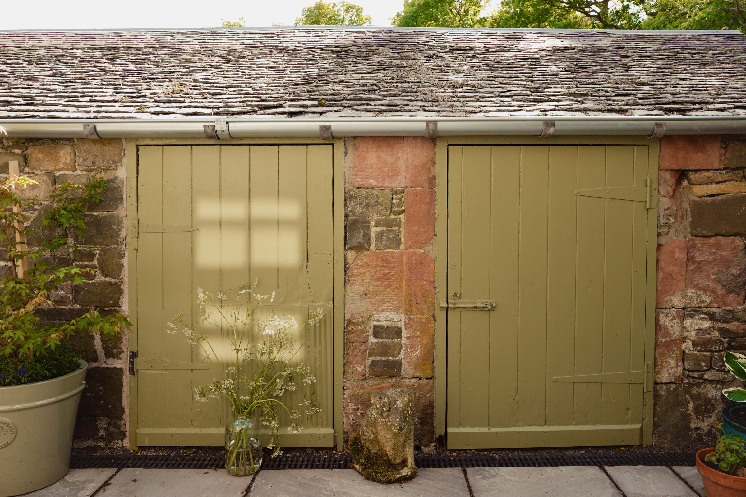 Two olive-green stable doors set in a red sandstone and rubble wall under a slate roof, a pot of cow parsley on the paving.
