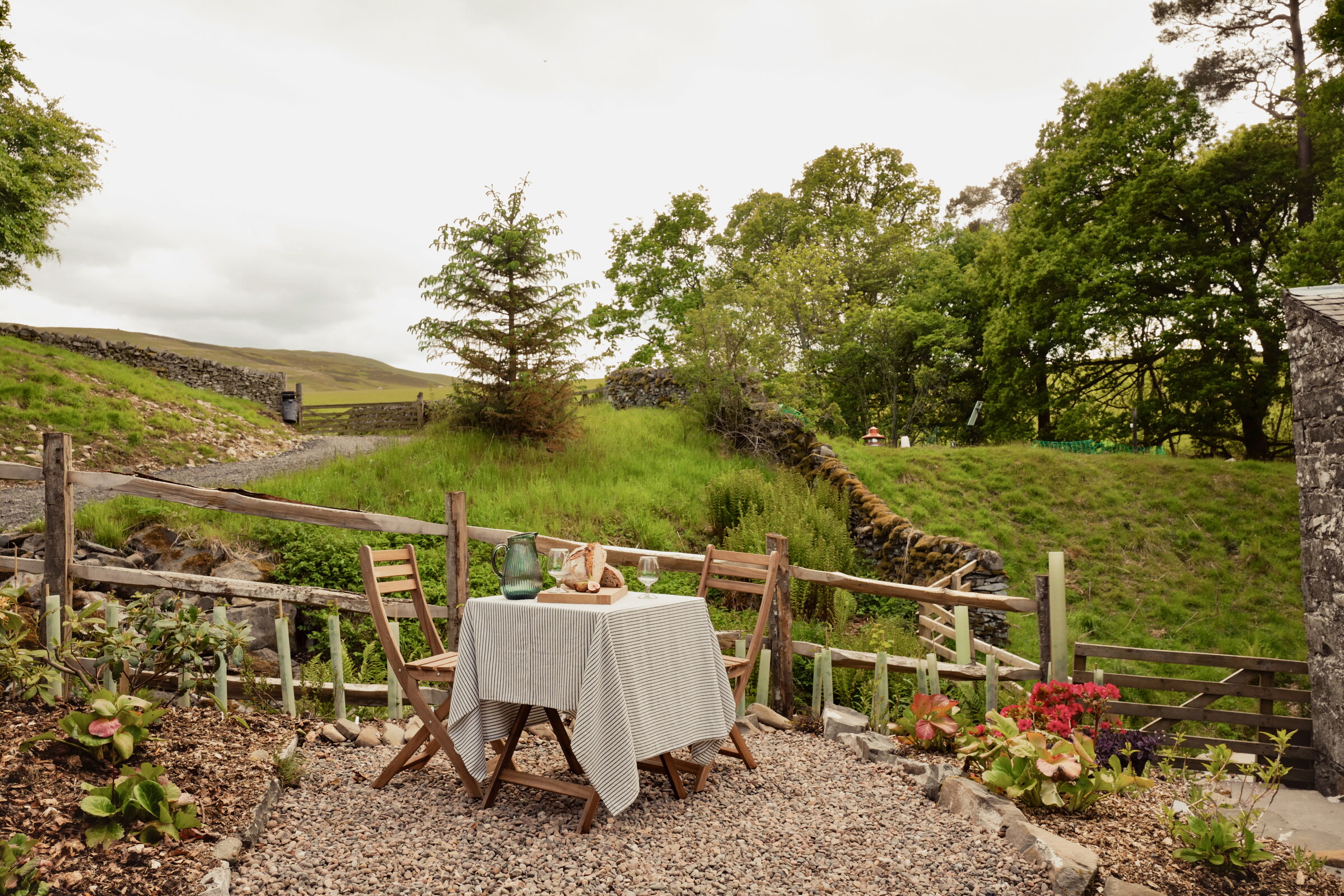 A table laid with a linen cloth and two chairs on gravel by a rustic fence above the Ladhope Burn, with trees and the hill behind.