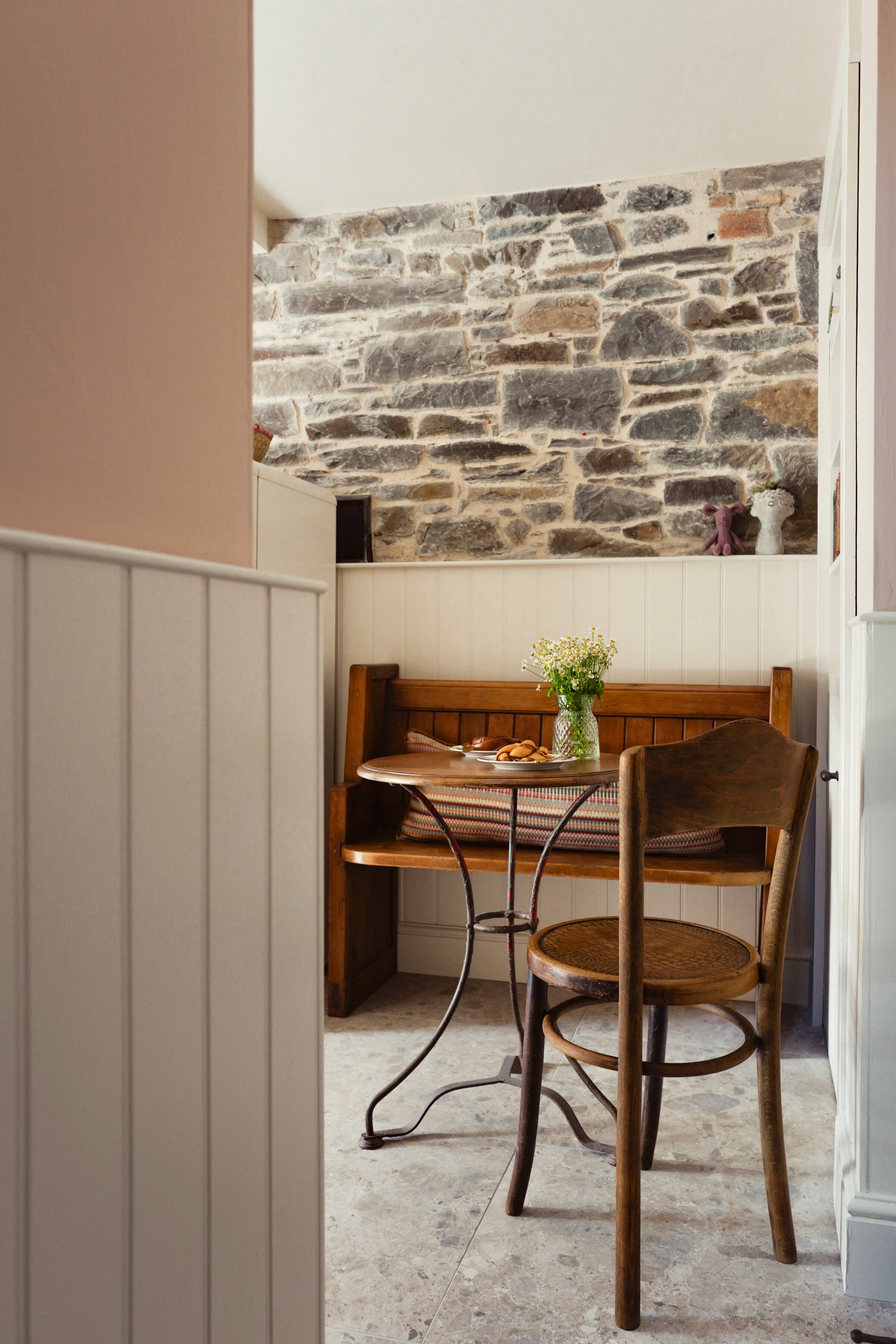 A pew and a bentwood chair at a little iron table under a strip of the scullery’s old stone wall, a jug of flowers on the table.