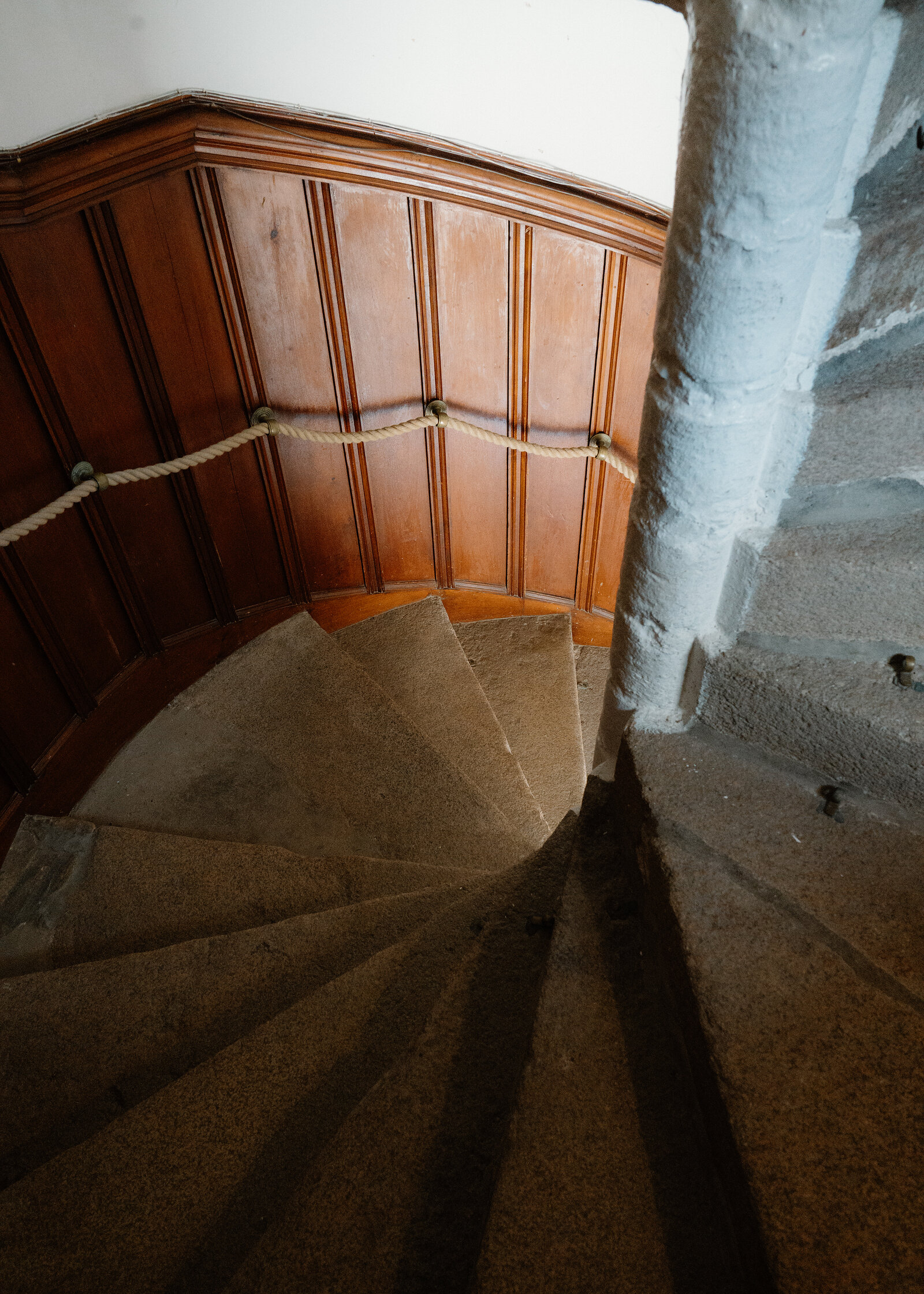 A stone spiral staircase descends inside Braemar Castle, with a rope handrail along wood-panelled walls and a whitewashed central pillar.