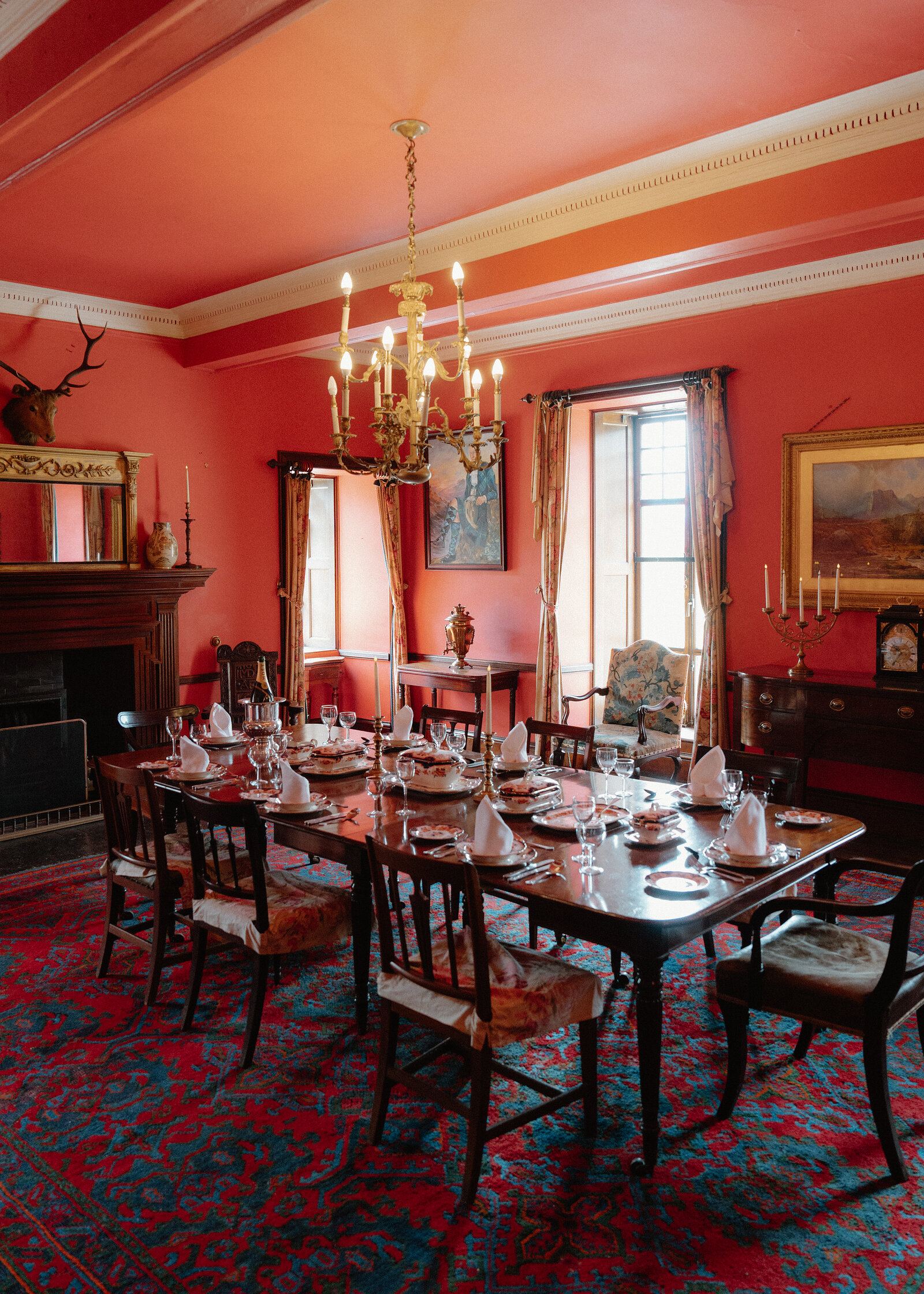 Formal dining room at Braemar Castle with coral-pink walls, a long table laid for dinner, a brass chandelier and mounted stag head above the fireplace.