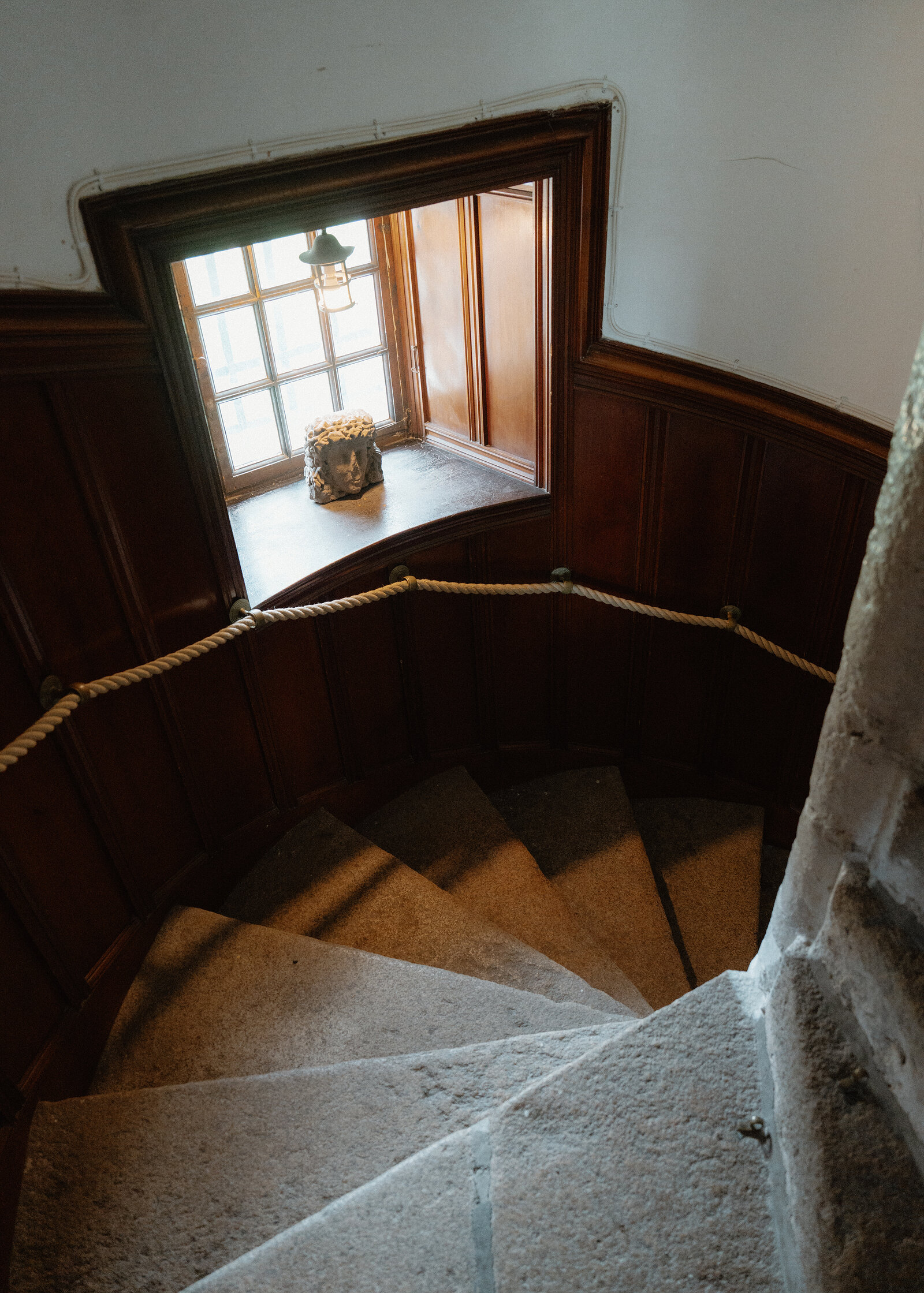 Looking down a stone spiral staircase with a rope handrail and wood-panelled walls towards a small window with a carved stone head on the sill.