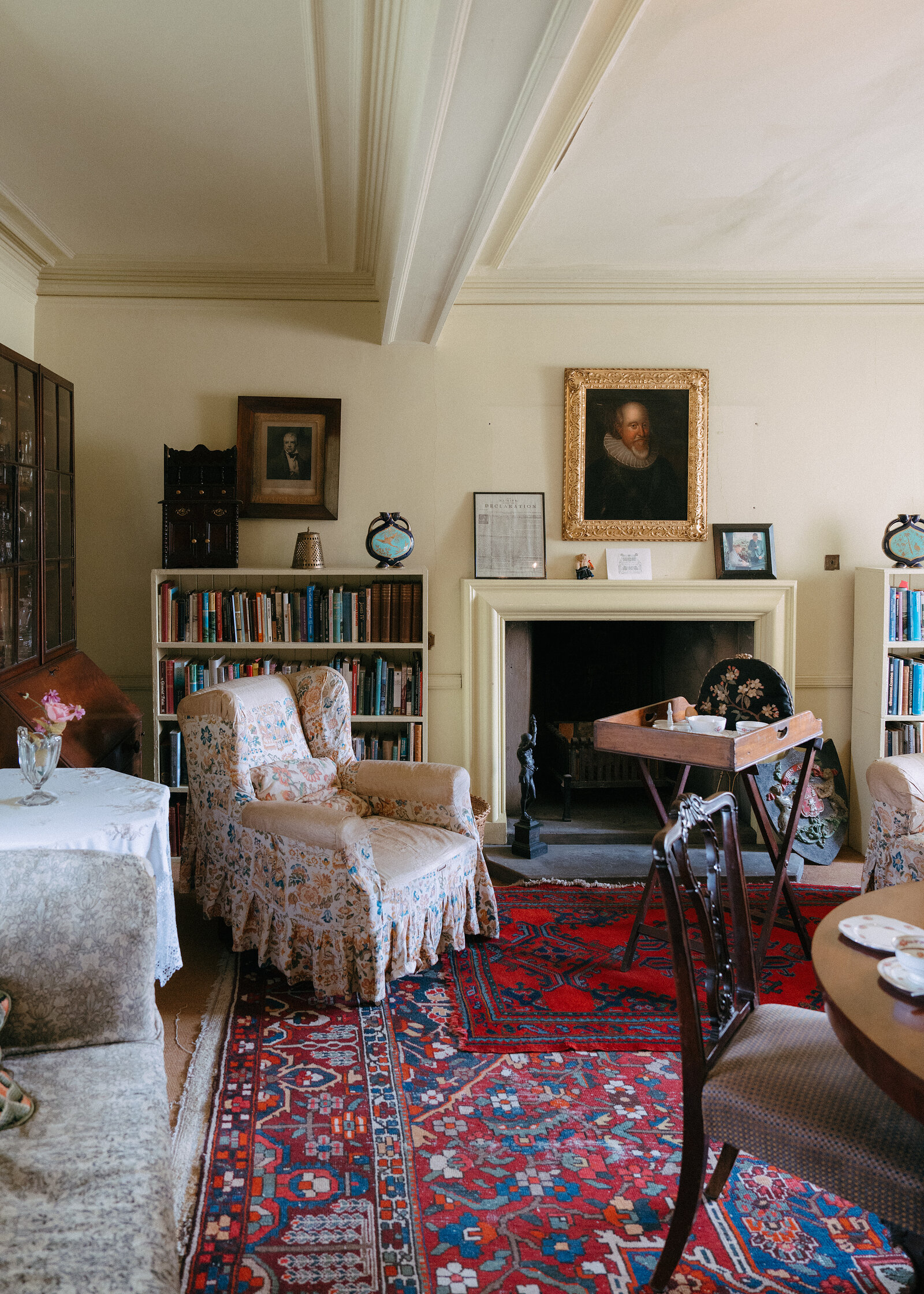 Sitting room with a floral armchair, red patterned rug, stone fireplace, bookshelves and a gilt-framed portrait above the mantel.