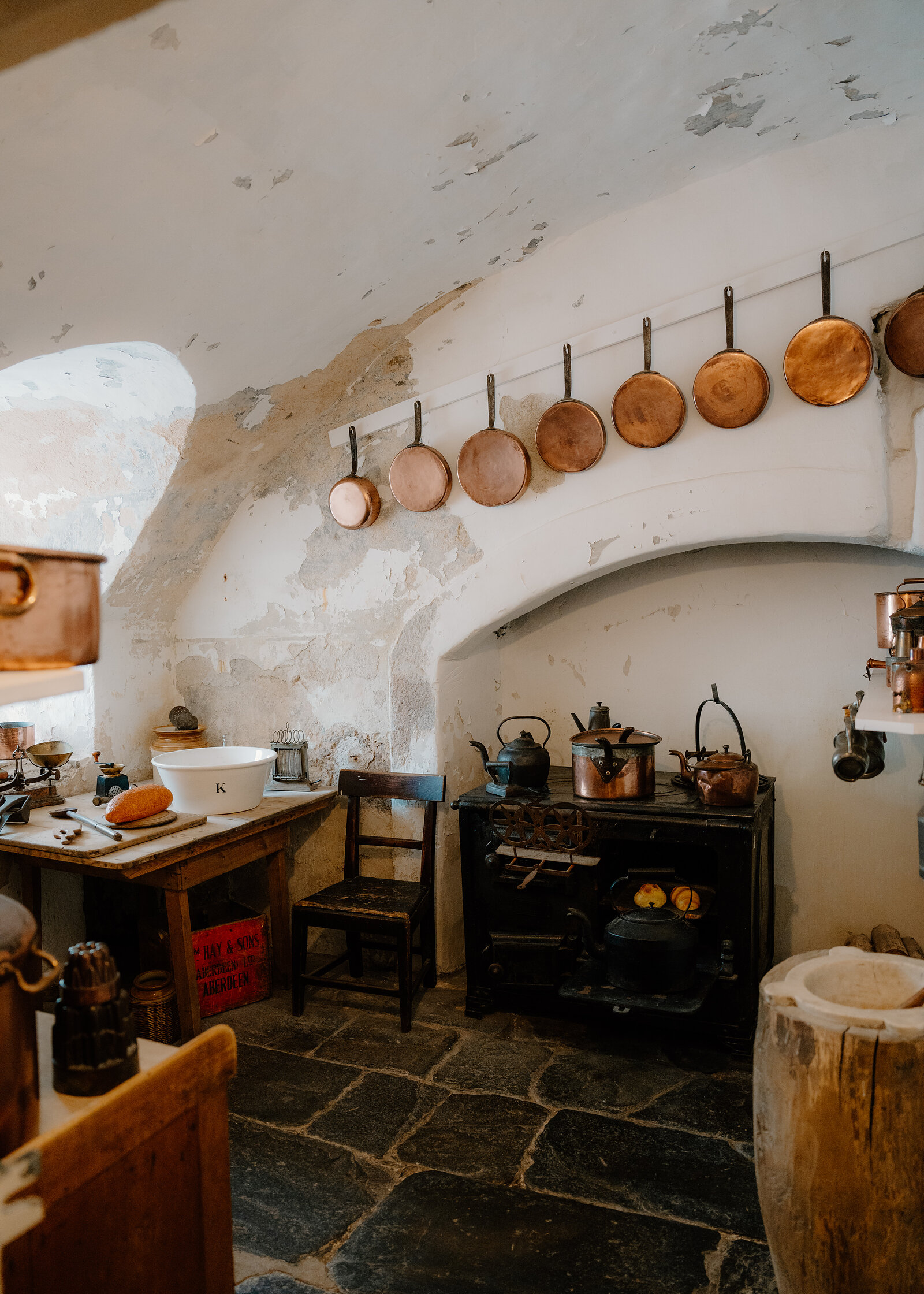 Old castle kitchen with copper pans hung along a whitewashed vaulted wall, a black cast-iron range, wooden chair and flagstone floor.