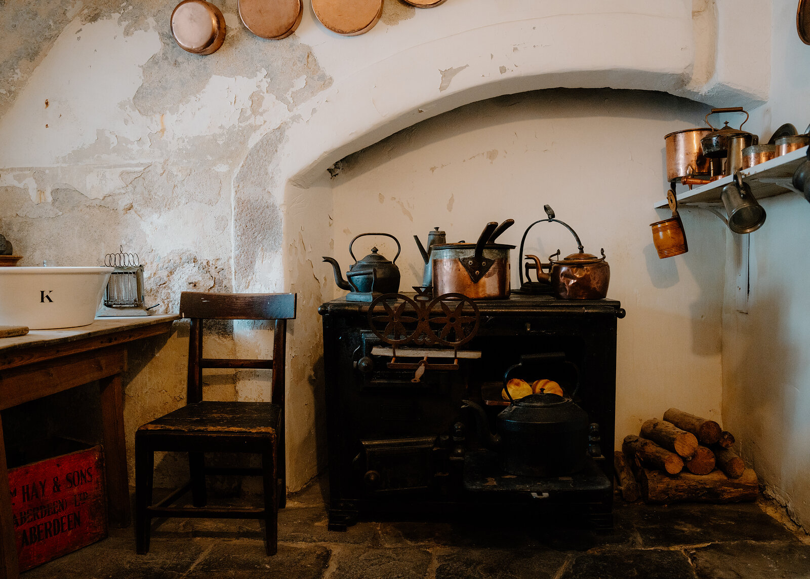 Old kitchen with a black cast-iron range, copper pots and kettles, a wooden chair and a stack of logs on a flagstone floor.