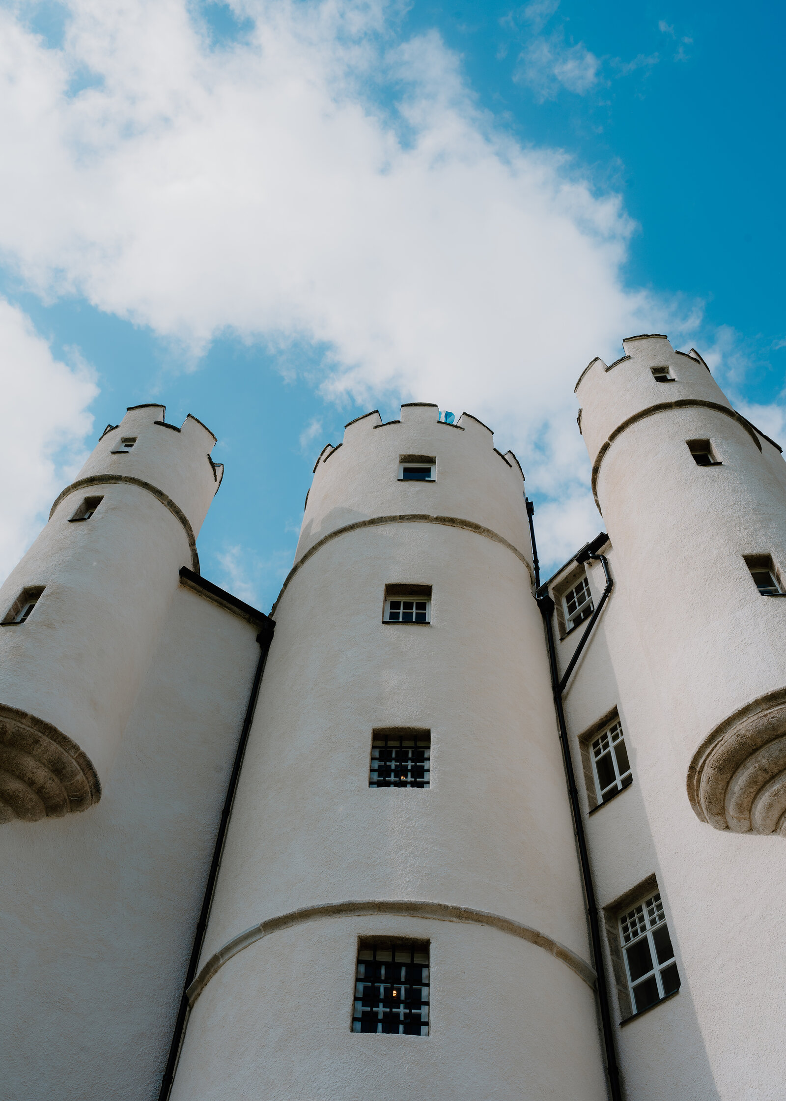 Braemar Castle's white harled round towers with crenellated tops, seen from below against a blue sky with white clouds.