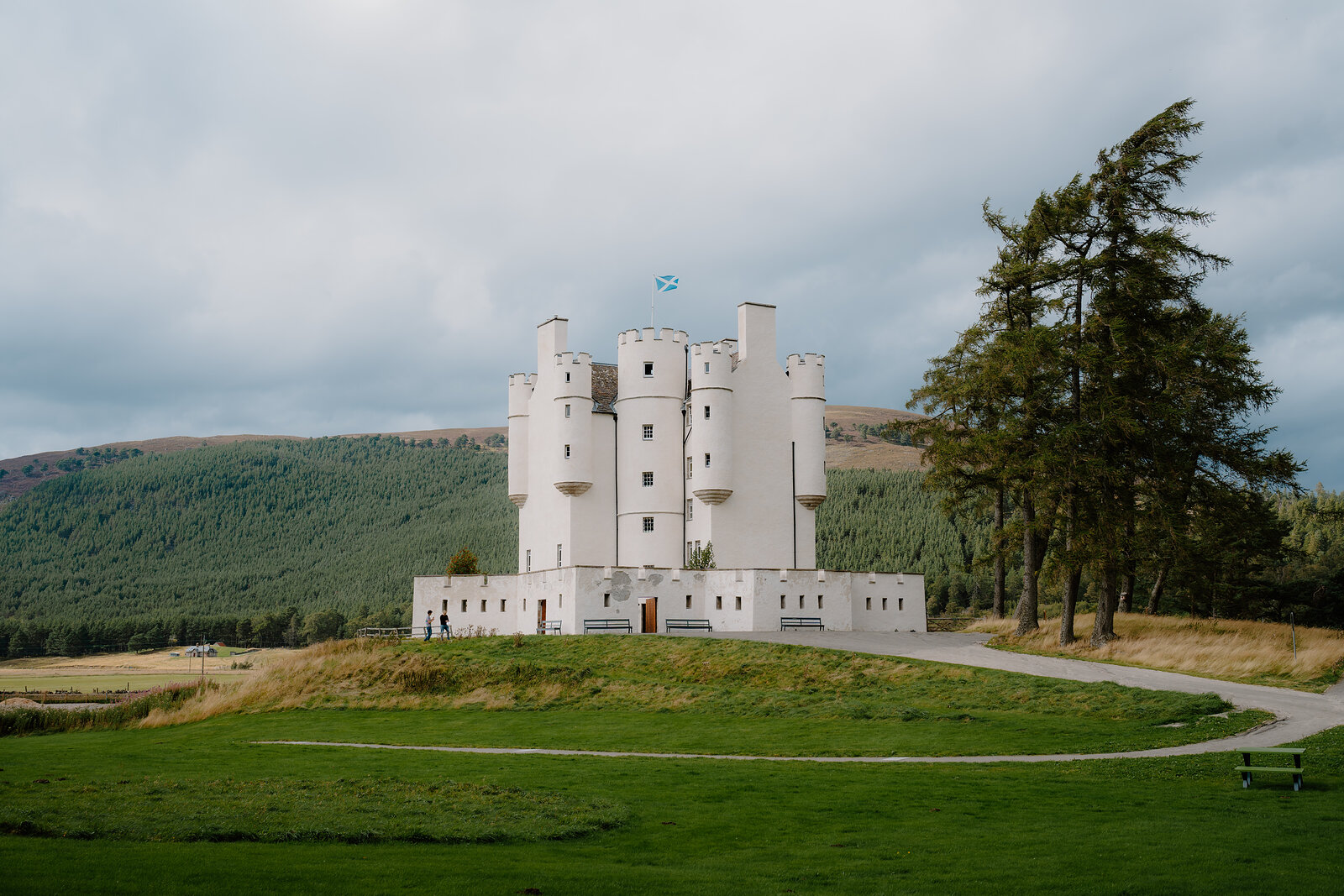 Braemar Castle, a white-harled tower house with turrets and a Saltire flying, stands on a grassy rise beside tall pines below a forested hill.