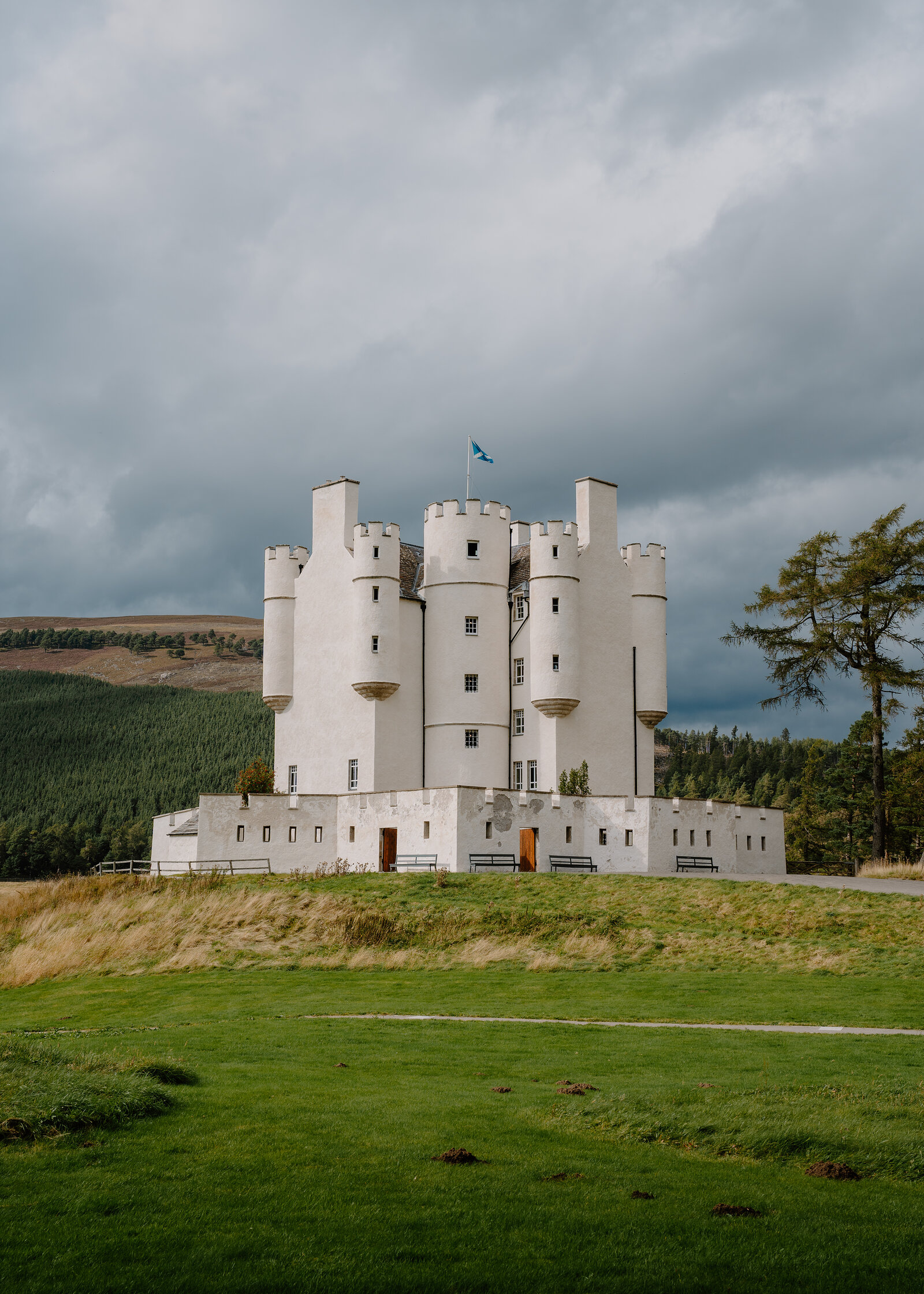Braemar Castle, a white-harled tower house with battlemented turrets and a Saltire flying, stands above a grassy slope under heavy grey cloud.