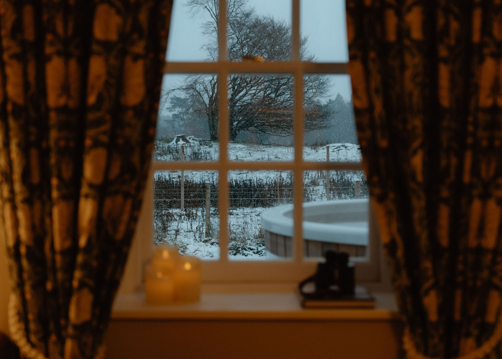 A landscape view through the window to the hot tub, the fence and a great bare tree in the snow, candles on the sill.