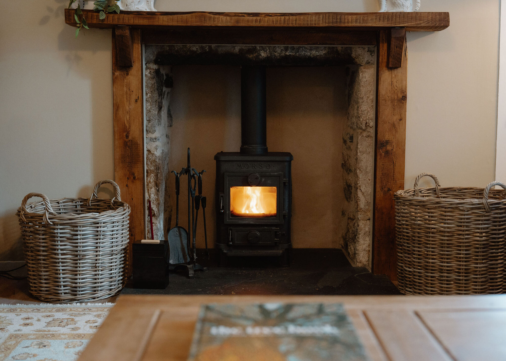 A landscape frame of the lit stove in its rough stone fireplace with baskets and fire irons.