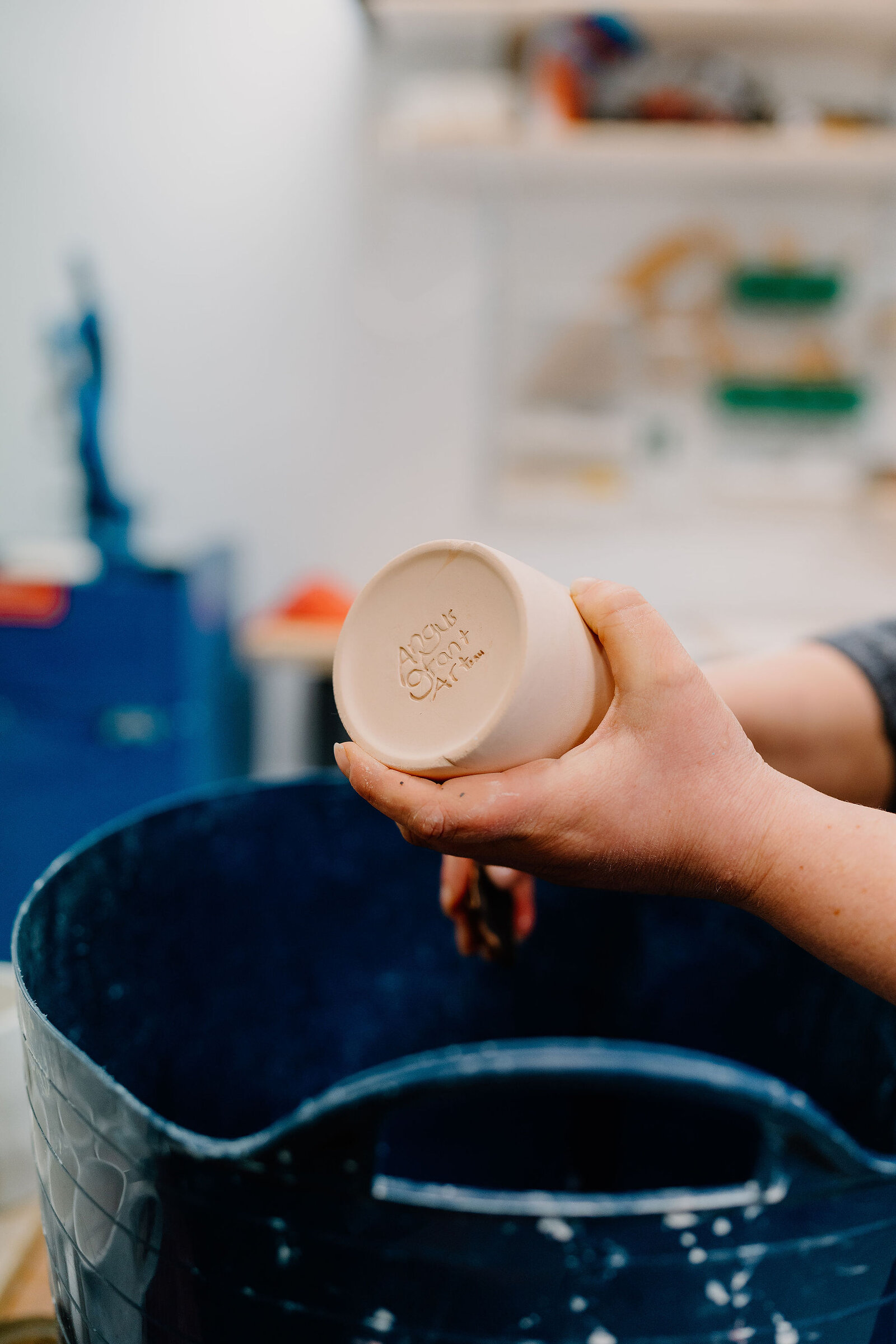A potter's hand holds an unfired stoneware beaker upside down to show the maker's stamp pressed into its base, a blue glaze bucket flecked with white below and workshop shelves out of focus behind.