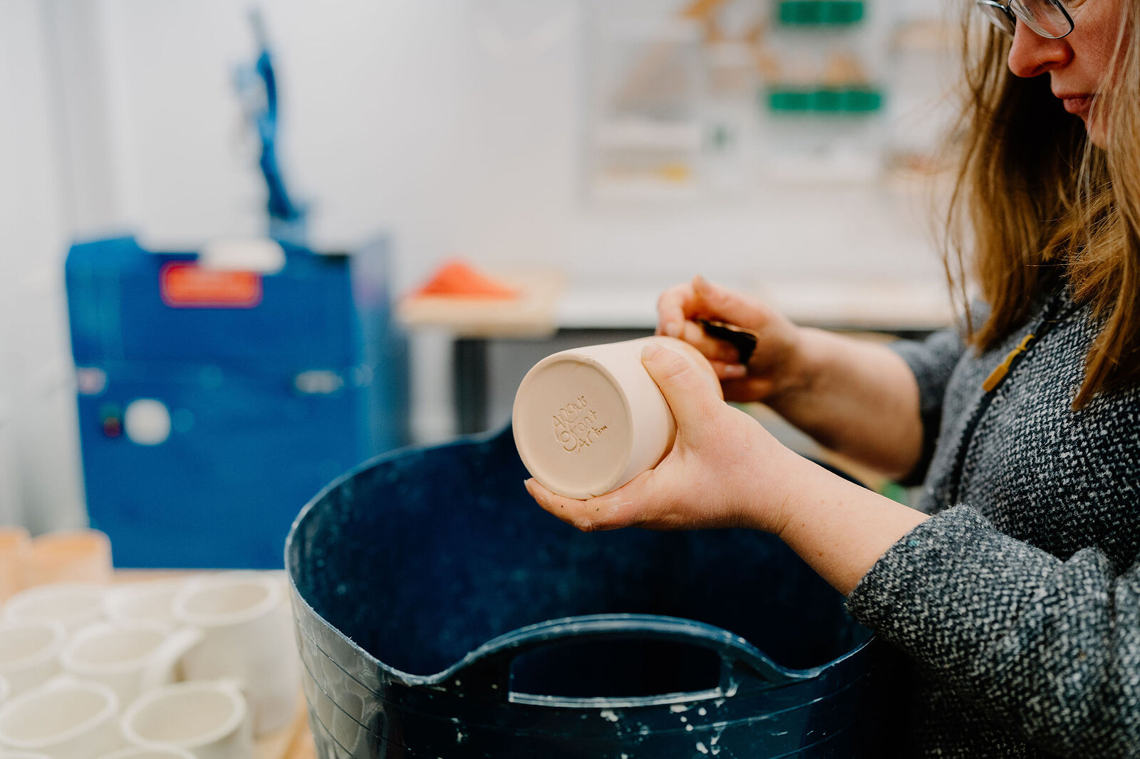 A potter's hands turn an unfired stoneware mug above a blue glaze bucket, tools and shelved pieces blurred in the workshop behind.