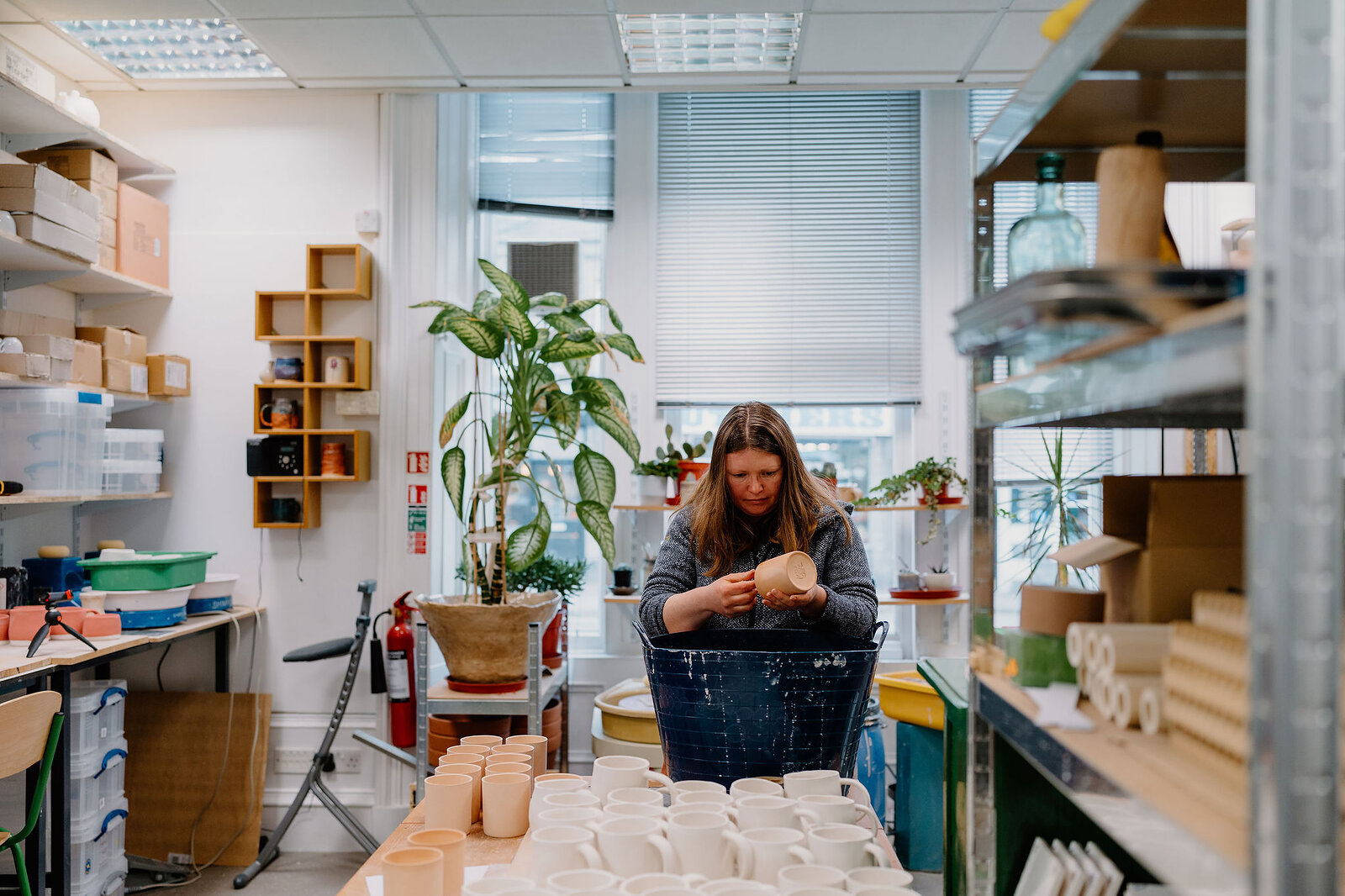 A potter looks down at a mug she is shaping, standing among shelving, a potted plant and a fire extinguisher in a bright studio.