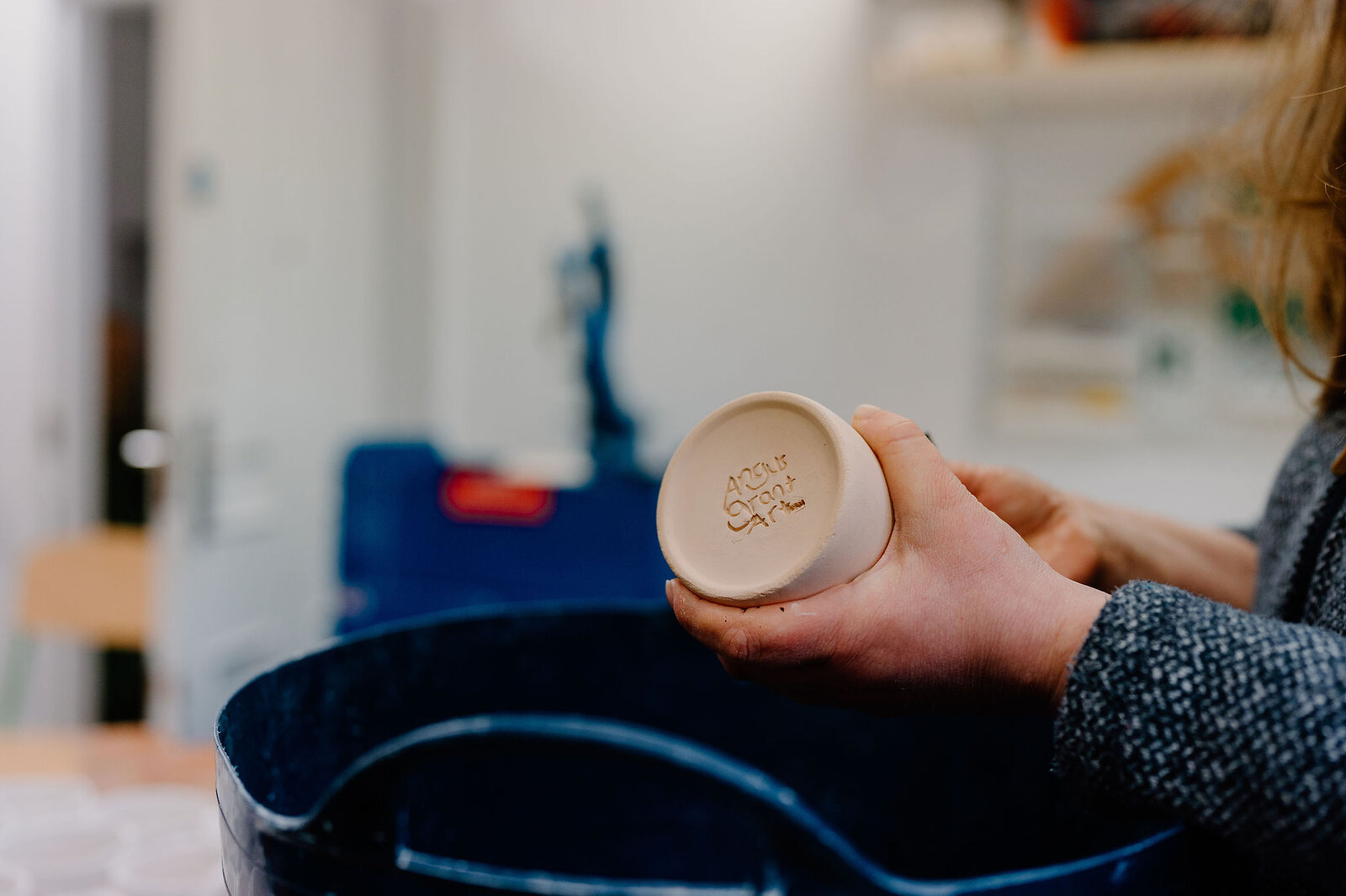 A hand holds a stoneware mug upside down to show a maker's stamp pressed into its base, a blue glaze bucket and workshop shelves blurred behind.