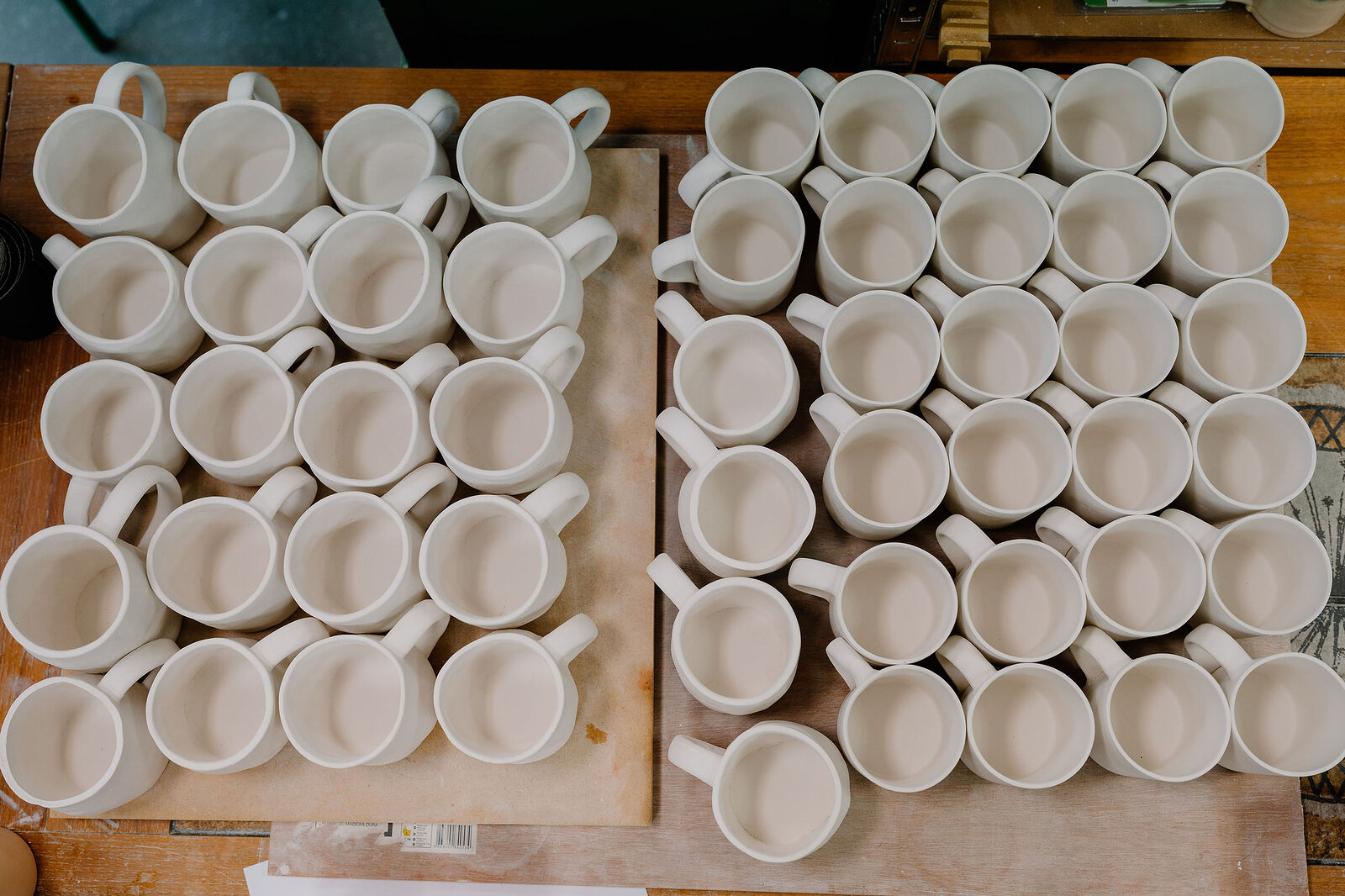 Rows of pale stoneware mugs stand drying on wooden boards, arranged in a grid across the workbench.