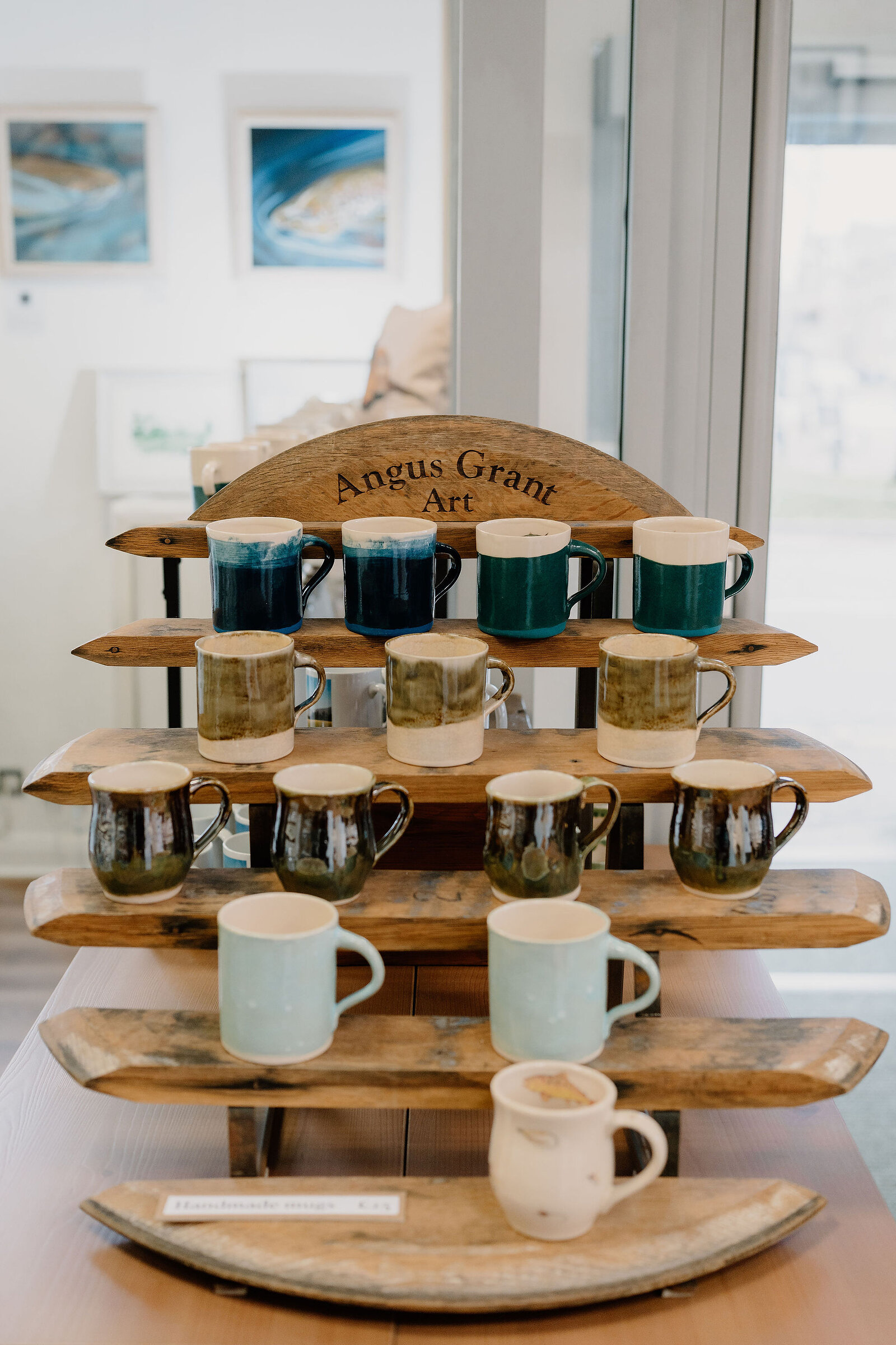 A tiered wooden stand branded Angus Grant Art displays rows of glazed mugs in blue, green and pale tones, framed art prints visible behind.